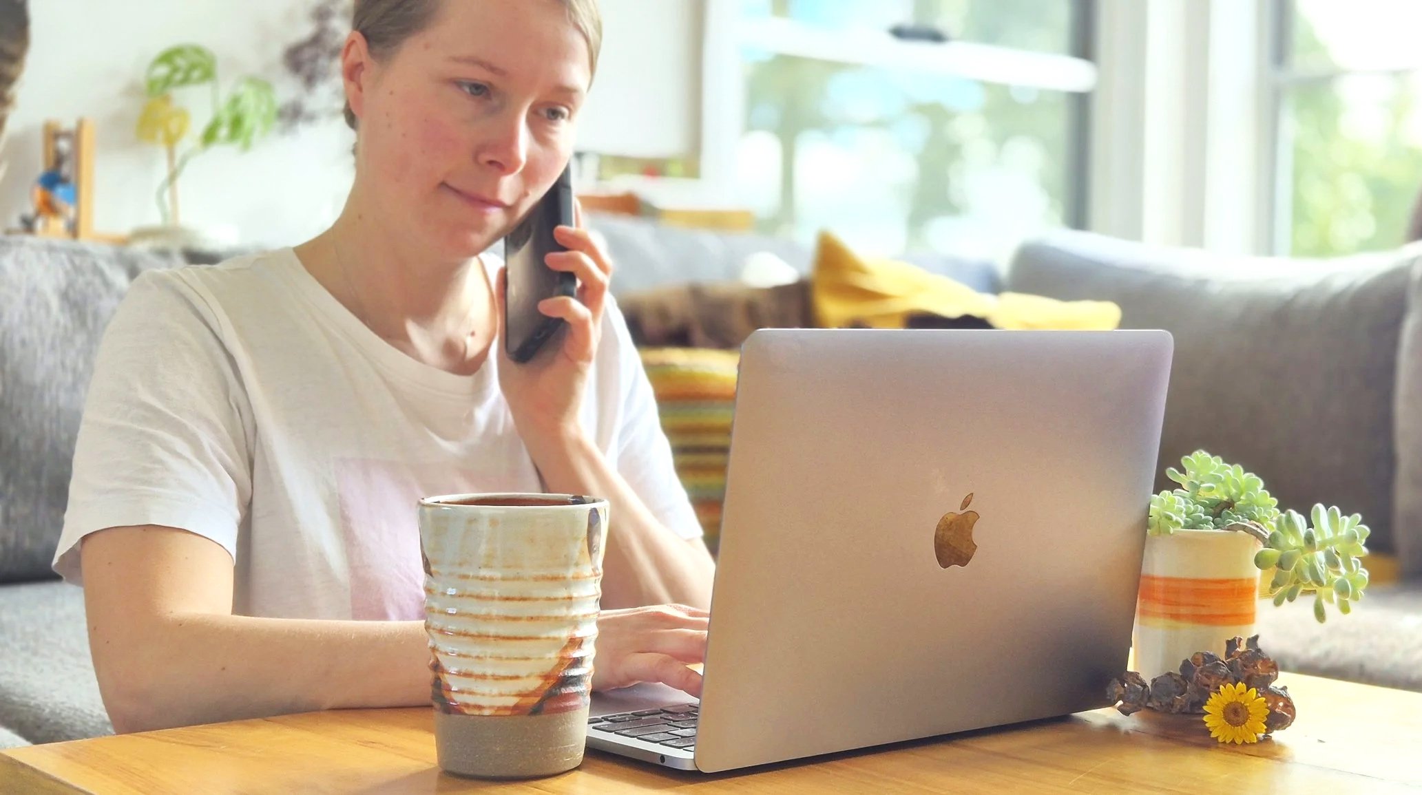 A young woman with short hair is sitting at a wooden table, talking on her mobile phone, with a laptop computer open in front of her. There is a cup of coffee and some succulent plants on the table, and a decorated mug in front of her. The background shows a bright living room with large windows and some cushions.