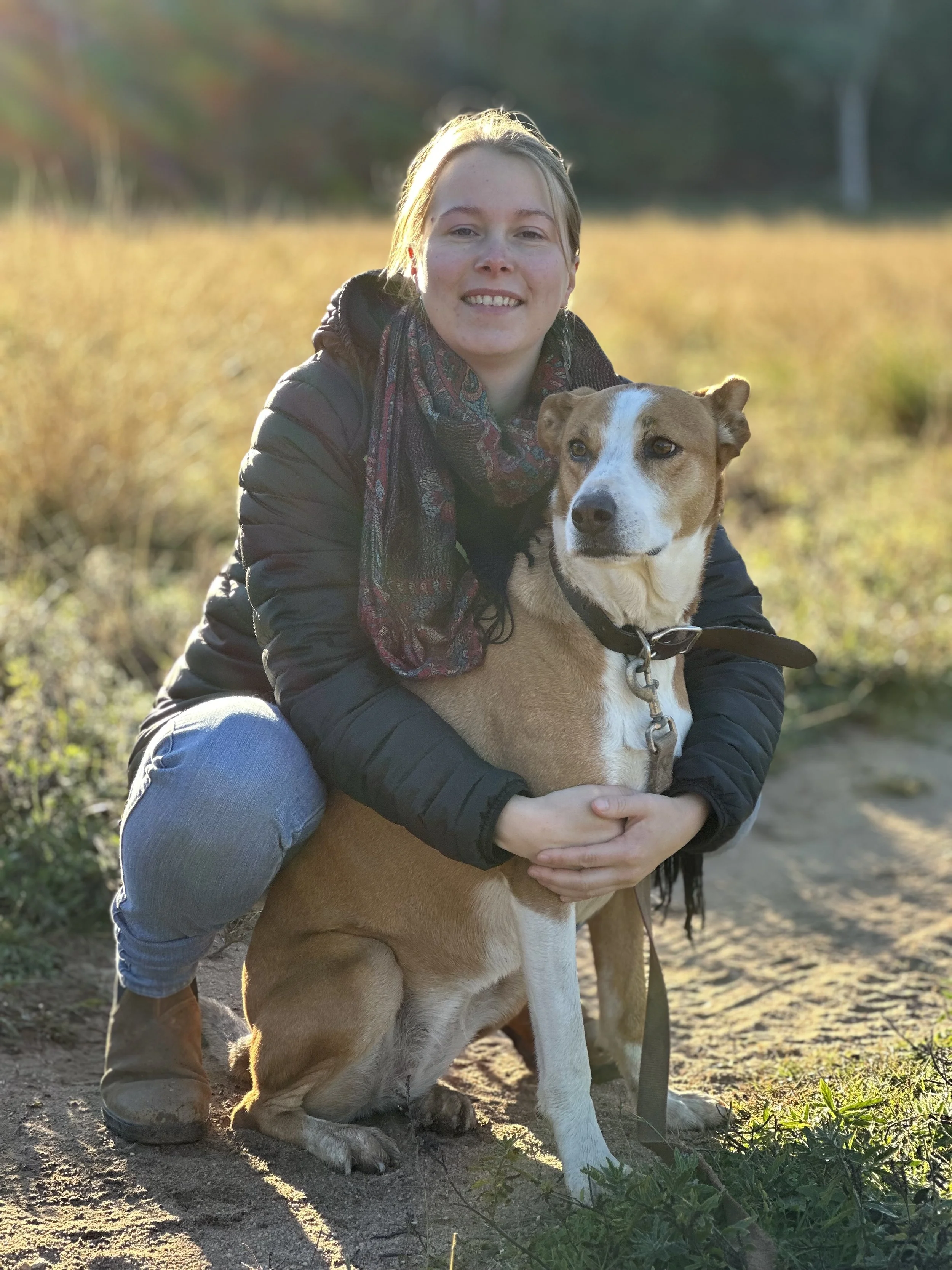 A woman with short light hair and freckles, dressed in a black jacket and multicolored scarf, smiling while hugging a tan and white mixed-breed dog with a white stripe on its face, outdoors in a sunny field with autumn trees in the background.