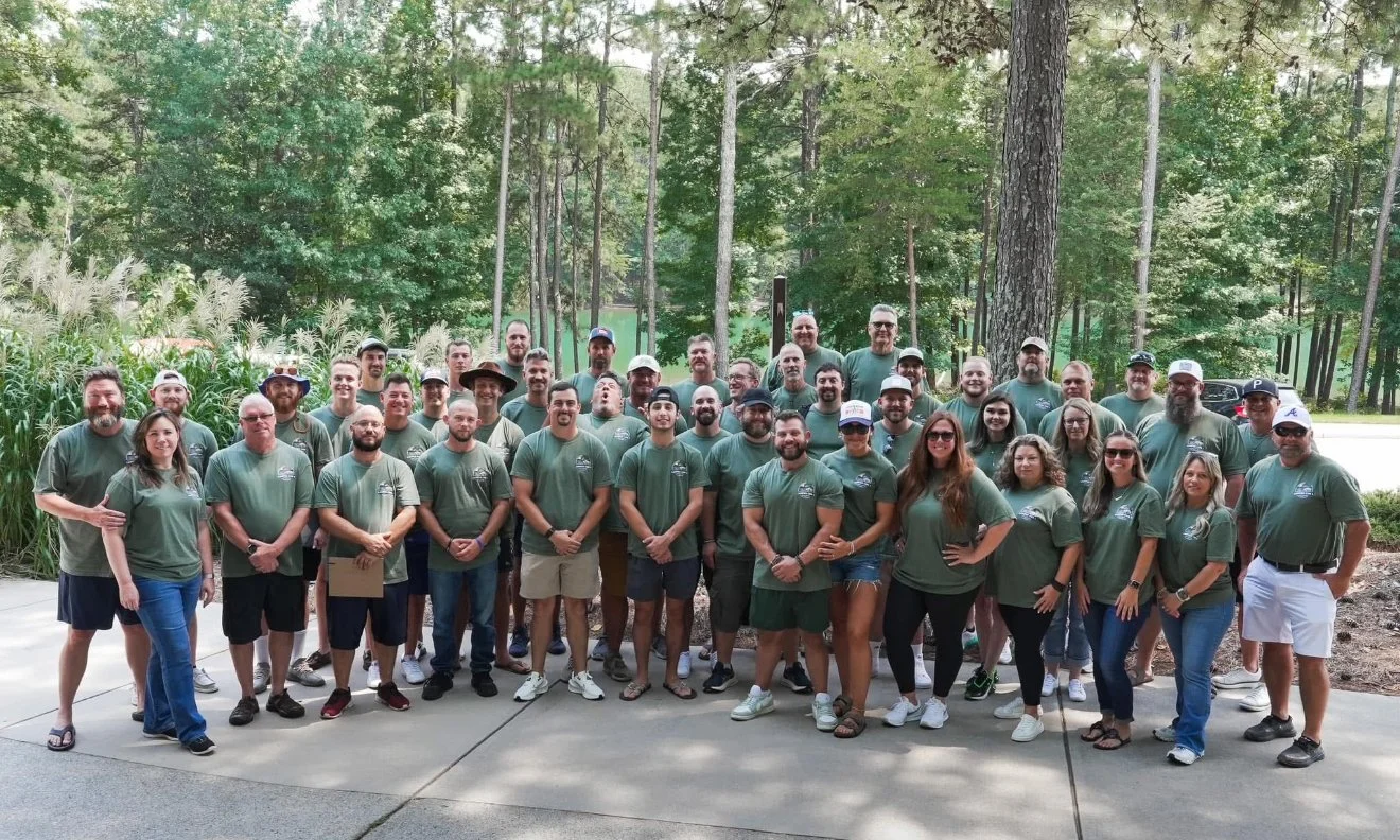 A large group of people wearing matching green t-shirts, standing outdoors on a concrete path surrounded by trees and greenery, posing for a group photo.