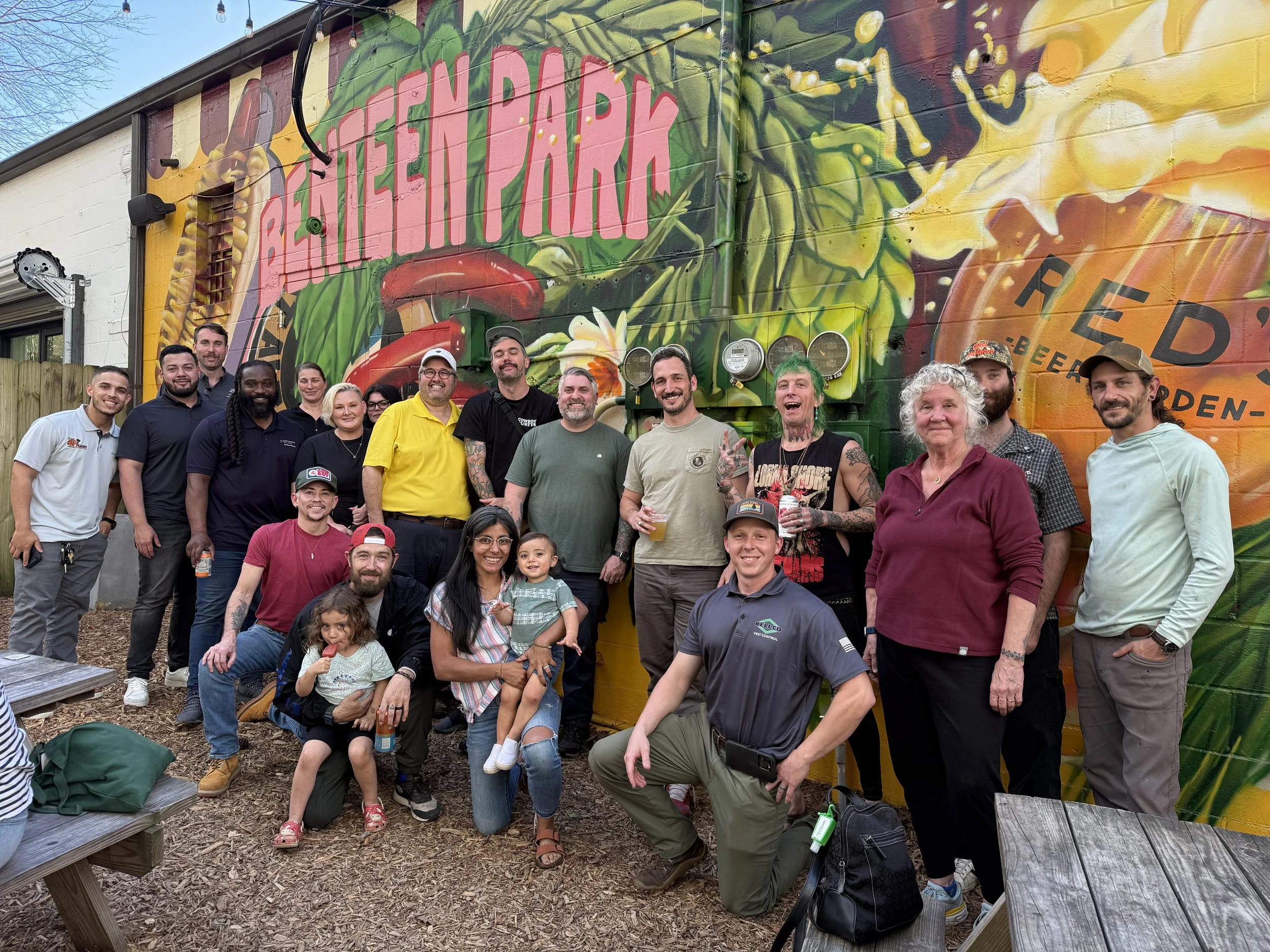 A group of diverse people, including men, women, and children, posing for a photo in front of a brightly painted mural that says 'BELL GENT FARM' with colorful illustrations of vegetables and plants. They are outdoors, and some are holding drinks, smiling, and making happy gestures.