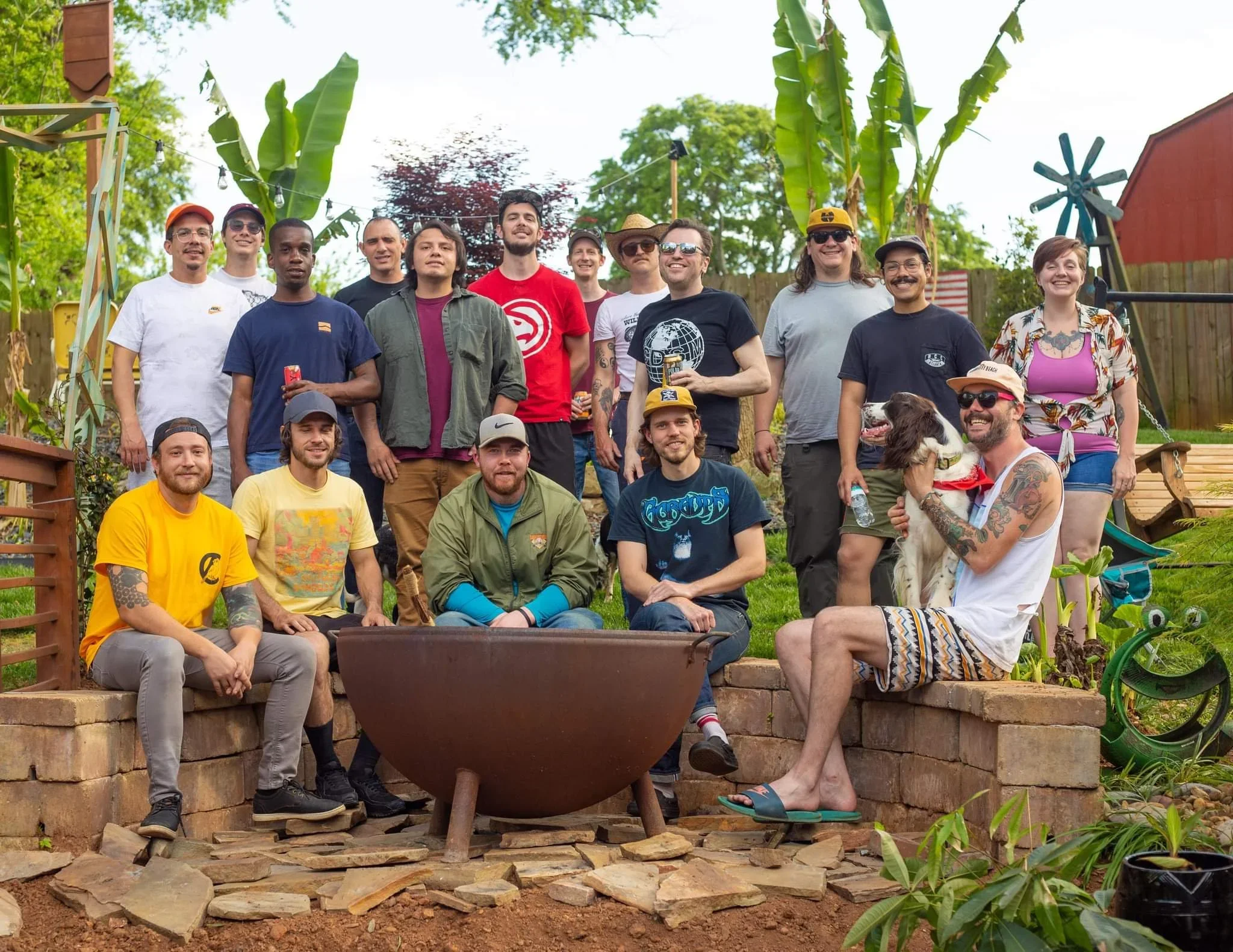 A diverse group of people gathered outdoors in a garden or yard, standing and sitting on a brick ledge, with greenery and trees in the background. They are smiling and appear to be enjoying a social event or celebration.