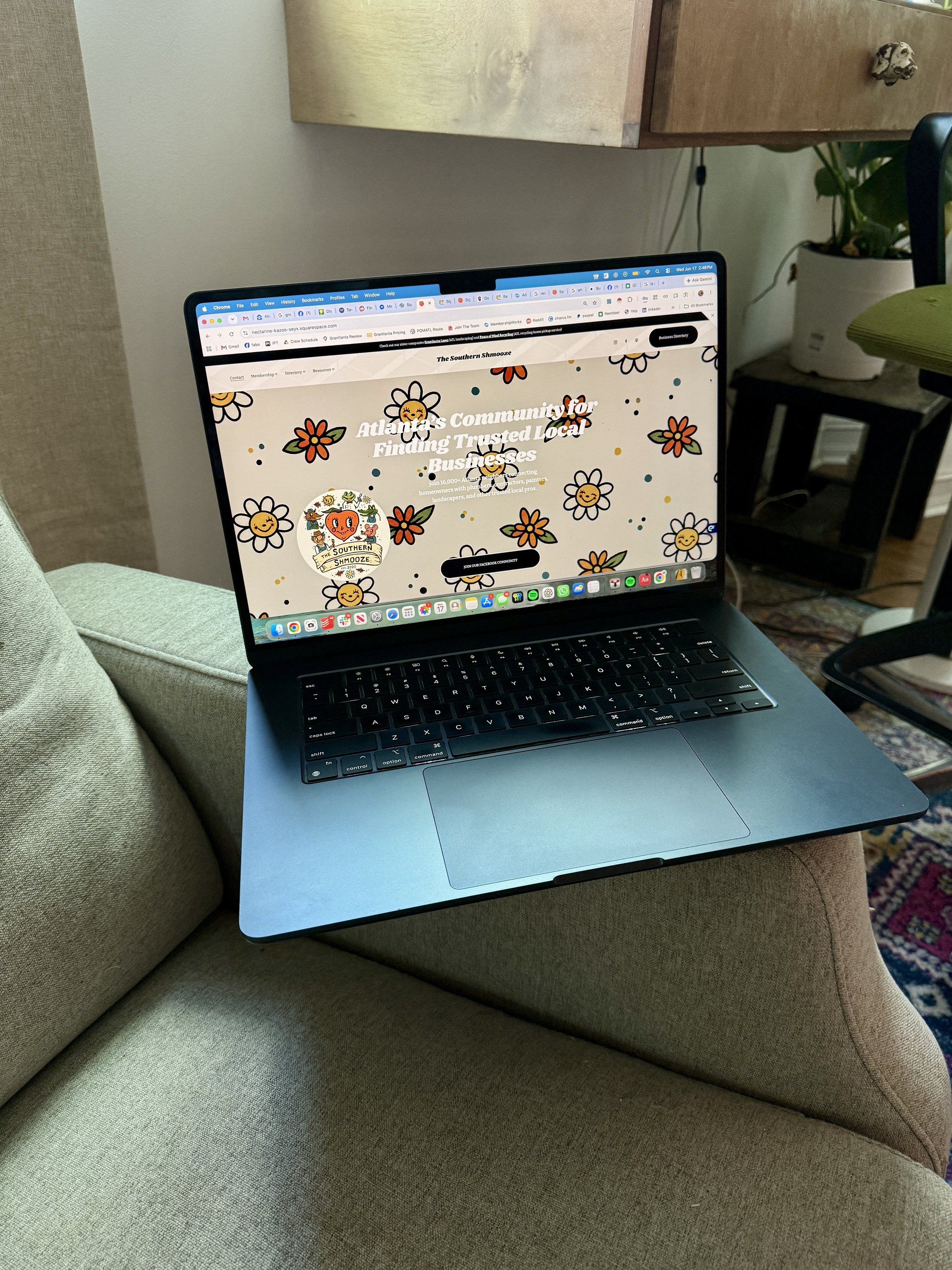 A laptop resting on a beige couch with a green cushion, surrounded by a wooden side table, a potted plant, and a patterned rug in the background.