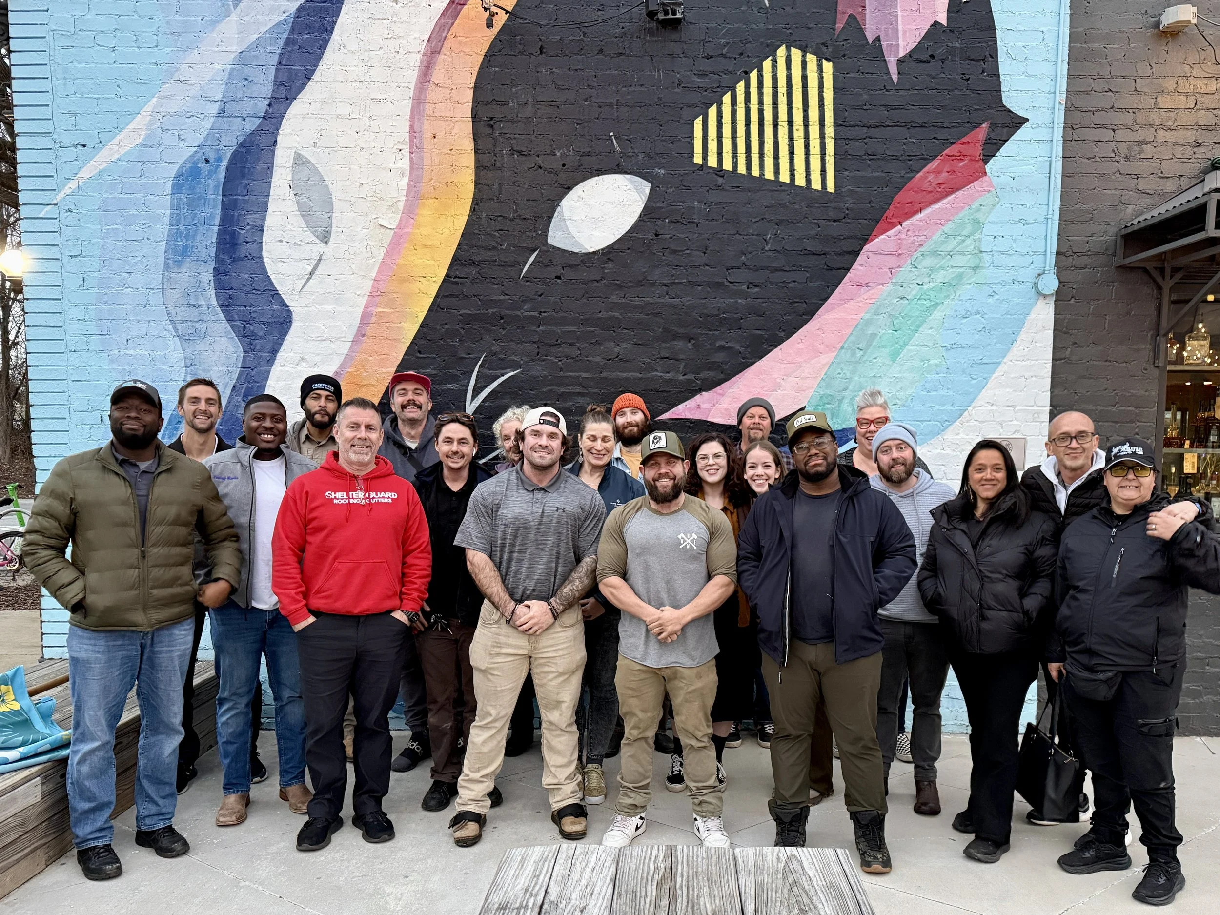 A group of diverse people posing outdoors in front of a colorful mural of a cartoon character with a rainbow mane and a black face. The mural is painted on a brick wall.
