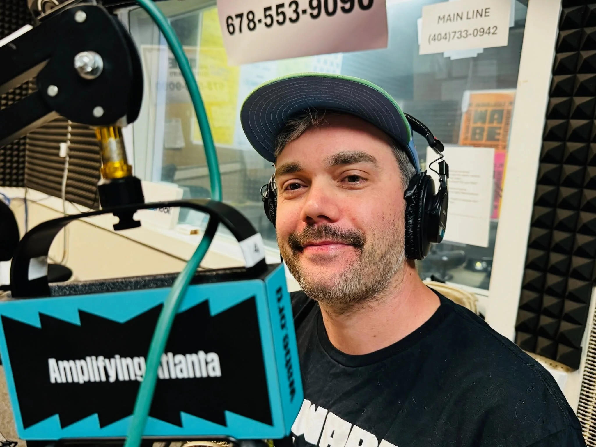A man wearing a green and black baseball cap and headphones taking a selfie in a recording studio with soundproof panels. There is a microphone with an arm in the foreground and various signs and posters on the glass window behind him, including a sign with a phone number and a sign reading 'Main Line' with a phone number.