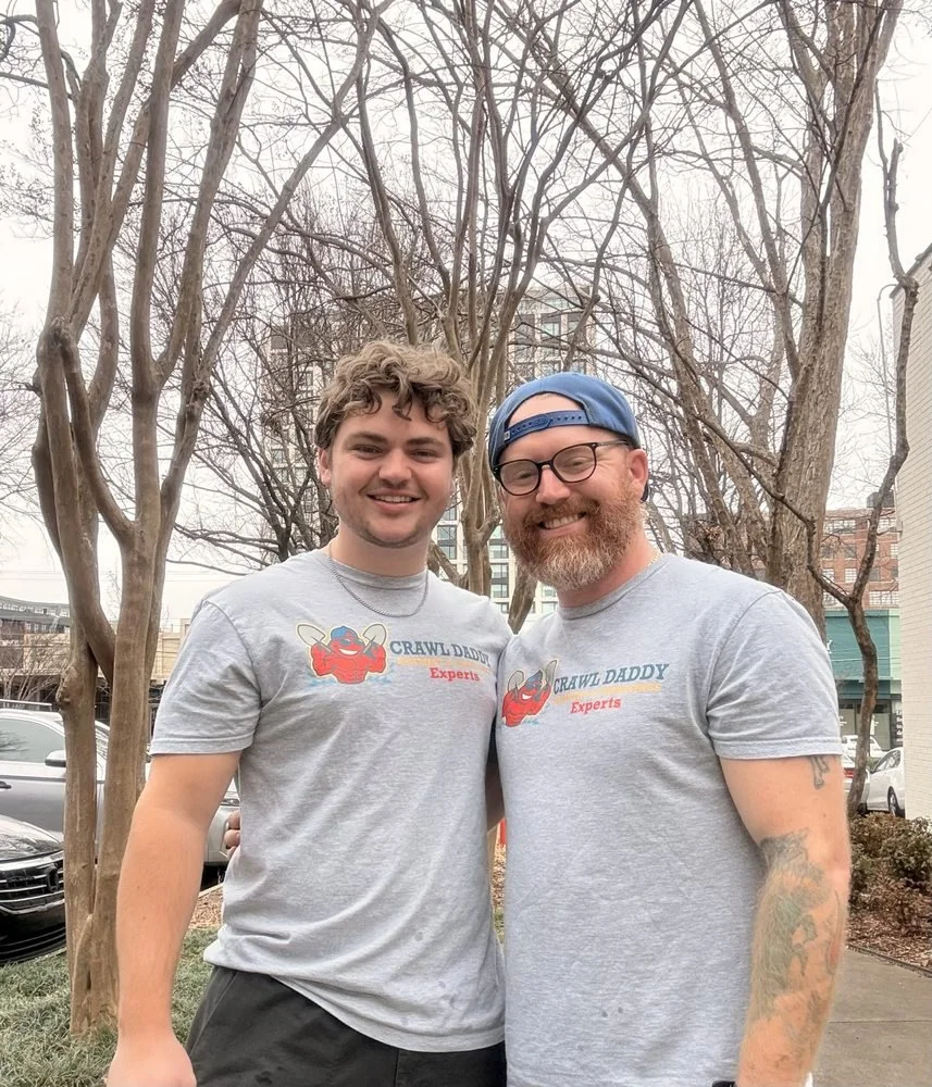Two men smiling, standing outdoors with leafless trees in the background. They are wearing matching grey T-shirts that say 'Crawl Daddy Experts' with a cartoon crab holding a spatula. One man has curly hair, and the other wears glasses and a blue cap.