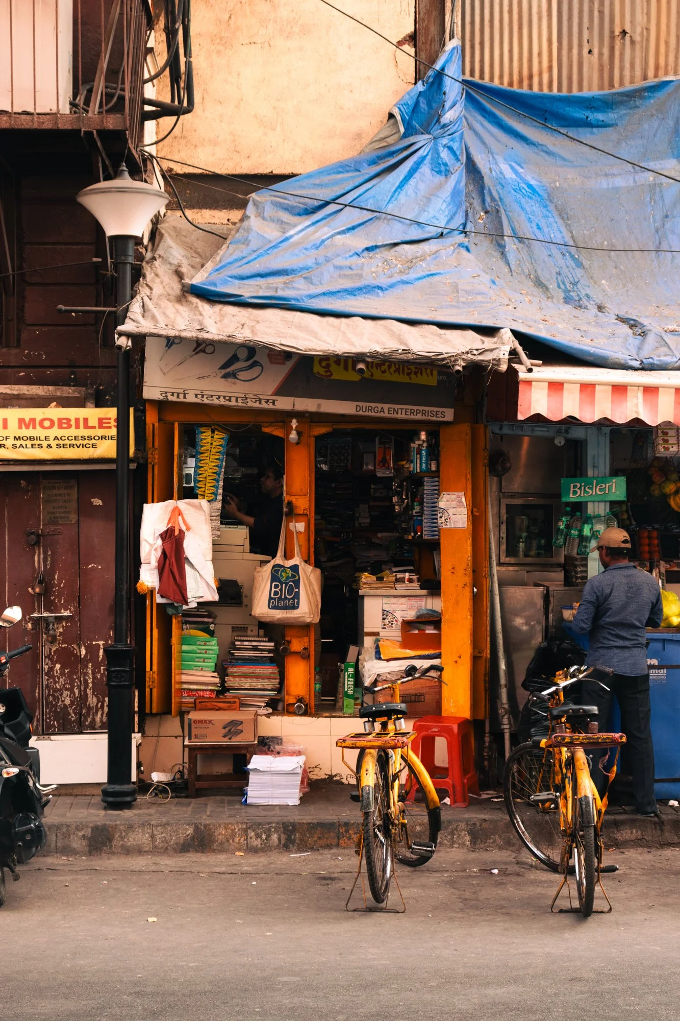 Street view of a small shop with a yellow entrance, books and goods displayed outside, bicycles parked in front, and a man standing near the shop. The shop name appears to be 'Durga Enterprises'.