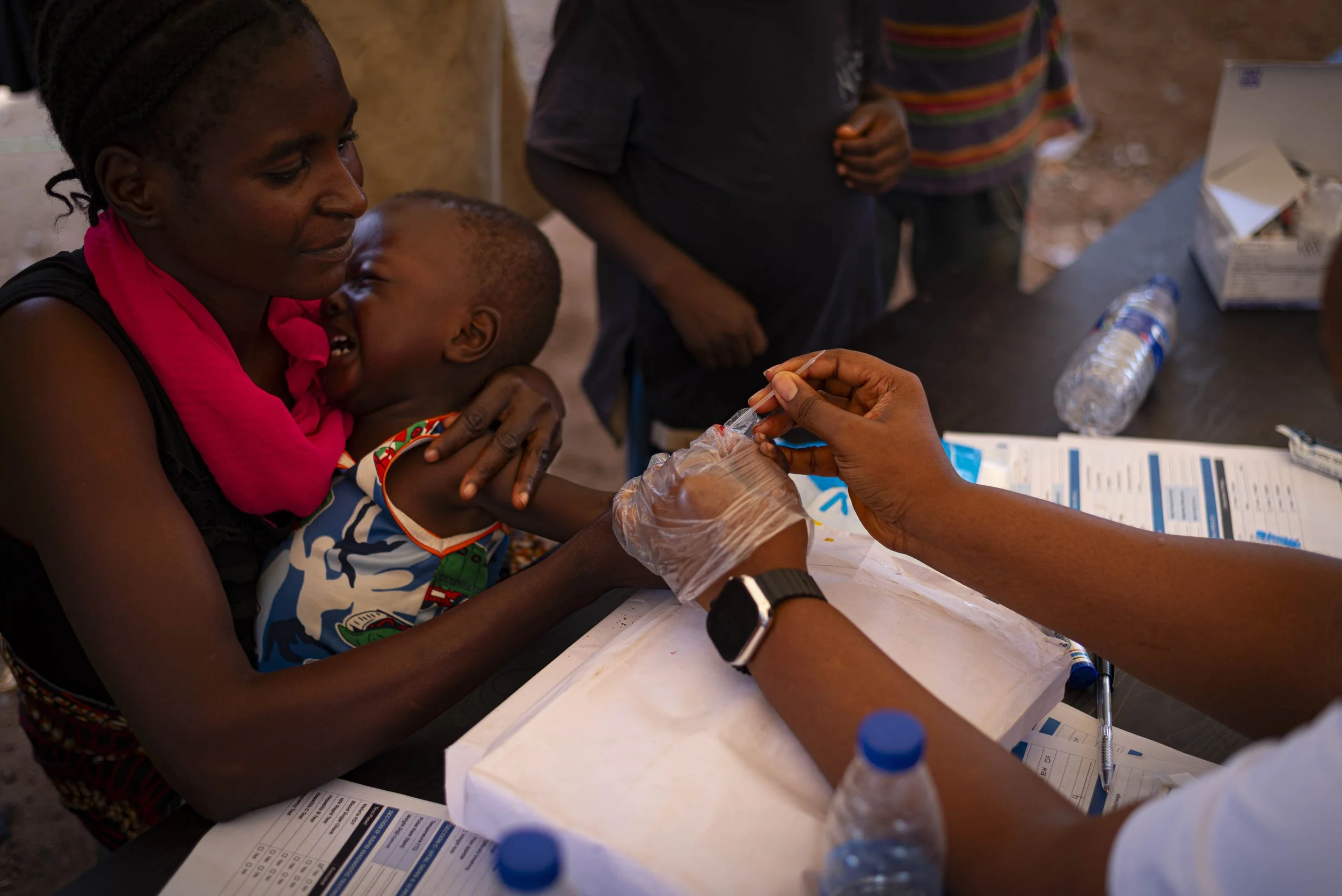 A woman holding a smiling child at a healthcare setting while a person administers a vaccination. The woman and child are engaged in a positive interaction, with medical supplies and informational materials on the table.