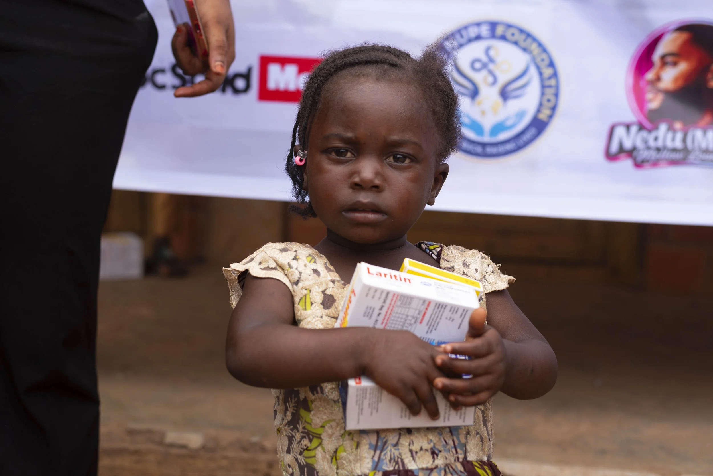 A young girl with braided hair holding a box of medication, standing outdoors with a banner featuring logos and a woman's face in the background.