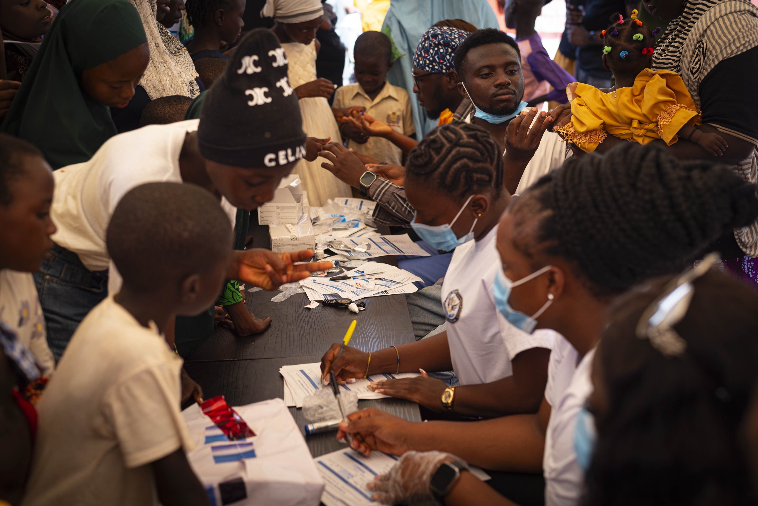 Group of people, including children and adults, gathered around a table at a health or community event, some wearing face masks, with various papers and medical supplies on the table.