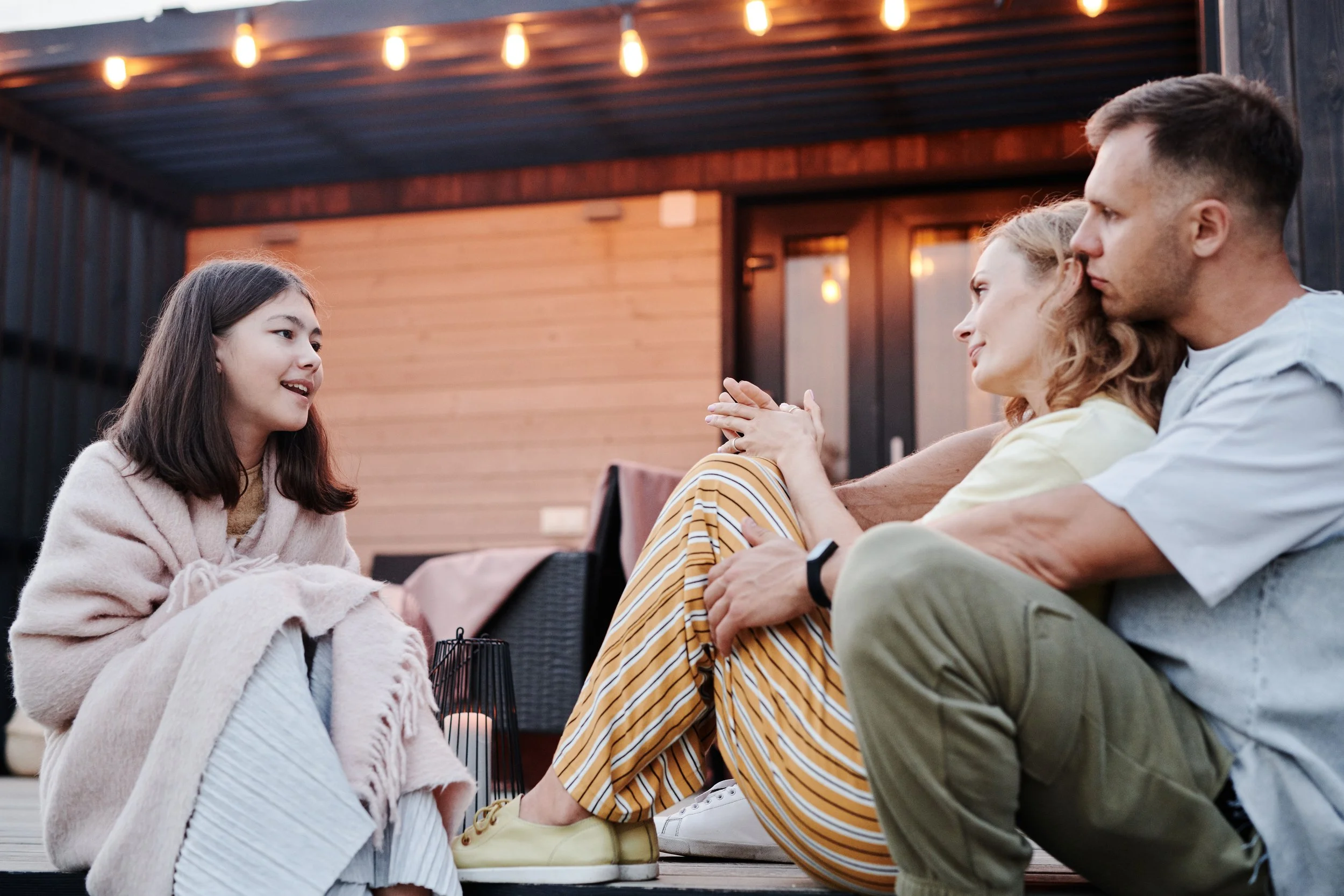 preteen girl, wrapped in a blanket, smiling, on the back porch talking to her parents