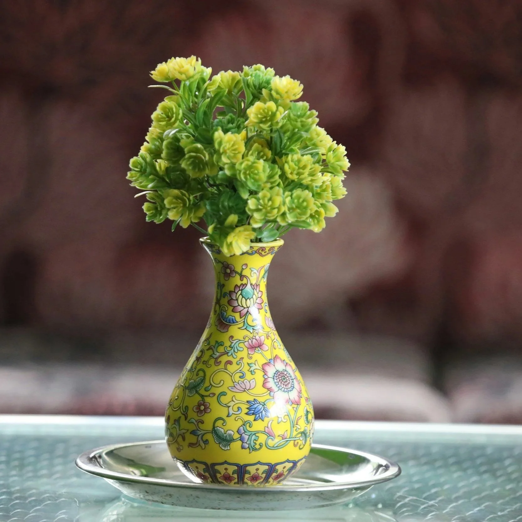 Yellow floral-patterned vase with small green and yellow flowers, placed on a glass saucer table.