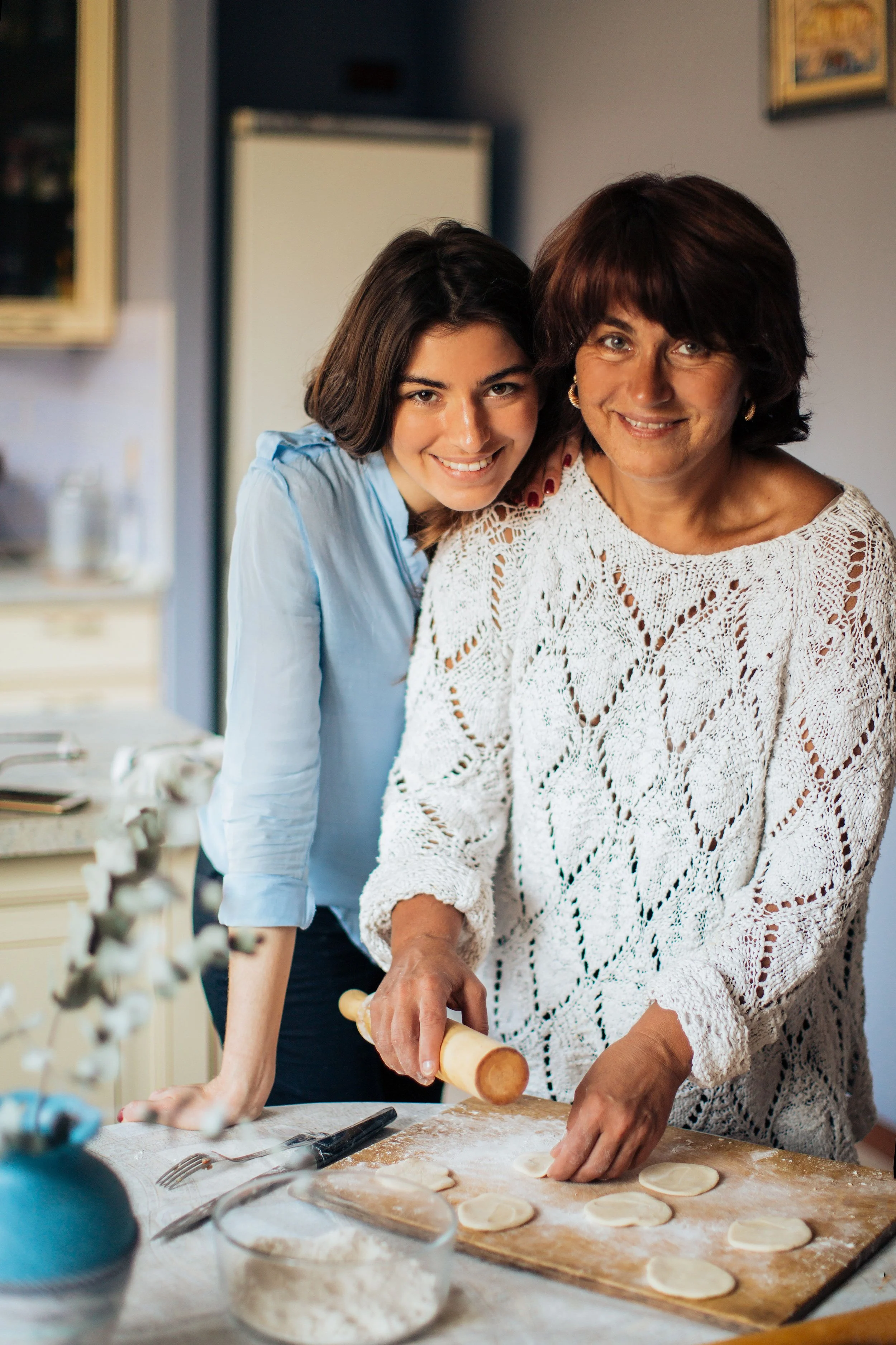 A woman and a girl are baking in a kitchen. The woman is using a rolling pin to flatten dough on a flour-dusted surface, while the girl stands beside her, smiling and watching. The kitchen has light-colored cabinets and a blurred background.