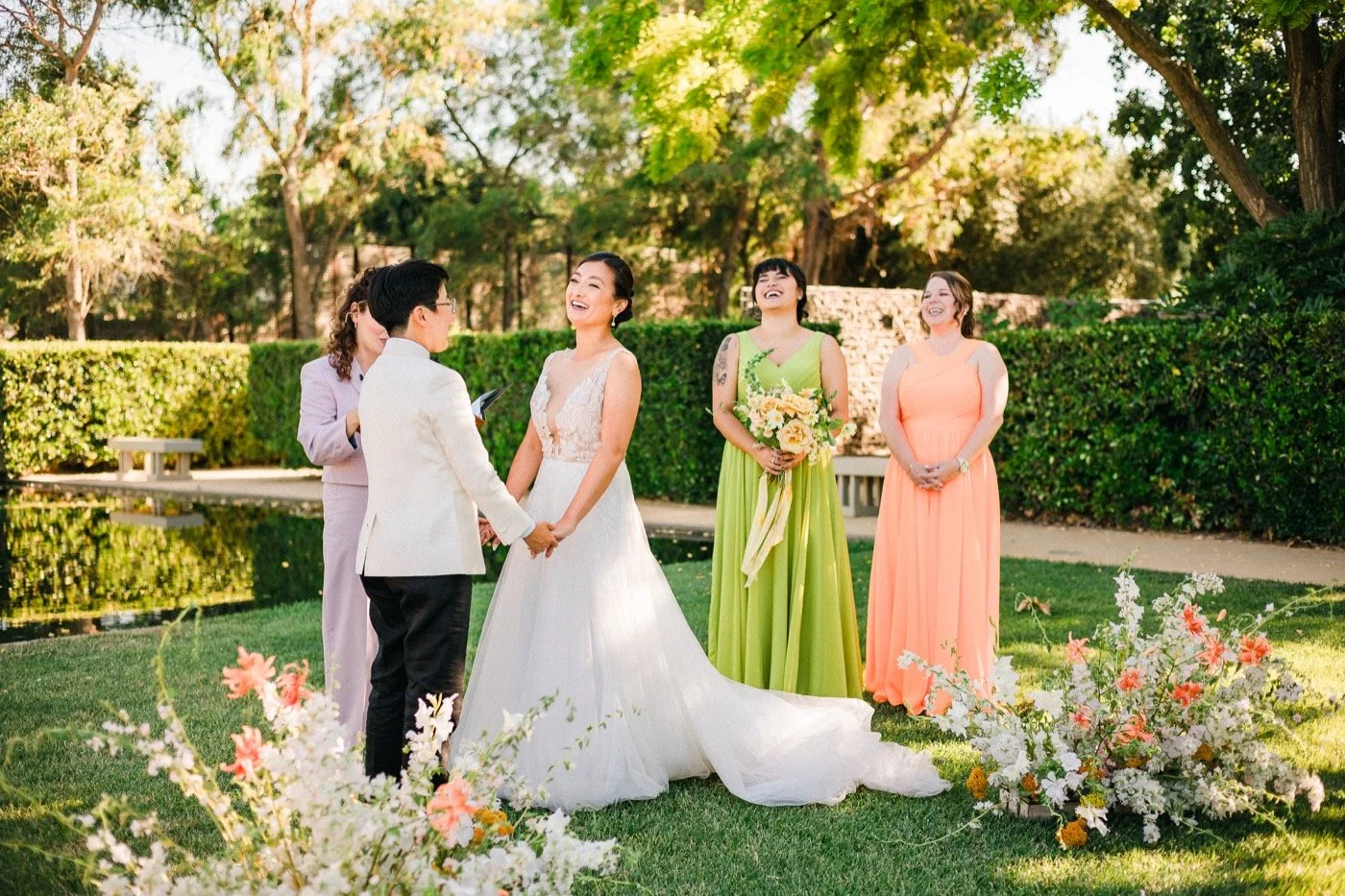 A wedding ceremony taking place outdoors with the bride and groom holding hands, surrounded by bridesmaids and a floral arrangement in a garden setting.