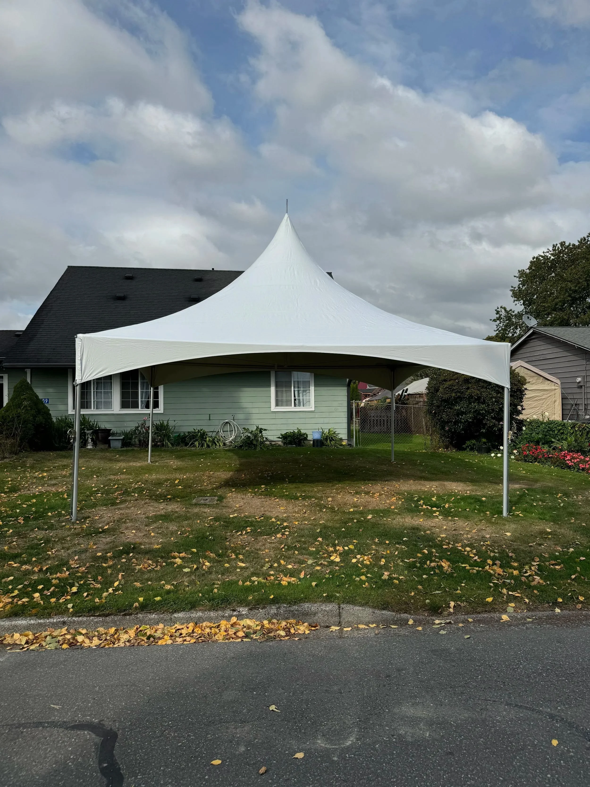 A white canopy tent set up in a residential backyard with greenery and houses around, and a cloudy sky overhead.