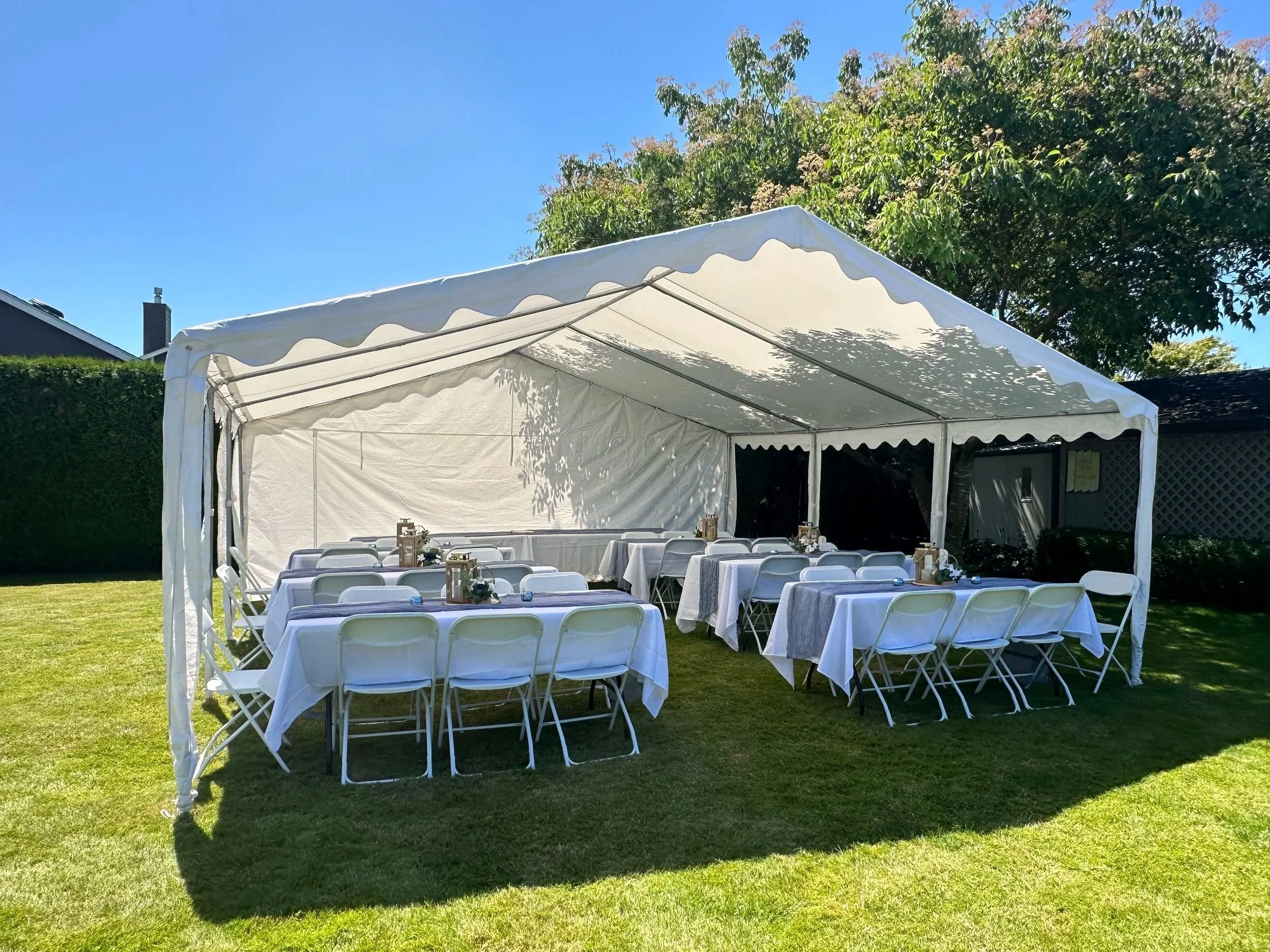 A large white event tent set up outdoors on a grassy lawn with tables and chairs underneath. The tent is surrounded by green bushes and trees, with a bright blue sky overhead.