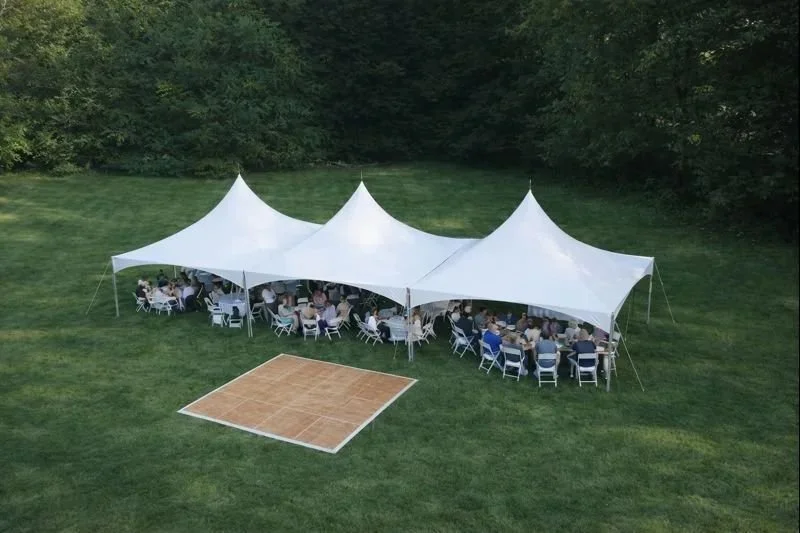 Outdoor event with tables under a large white tent in a grassy area, surrounded by trees.