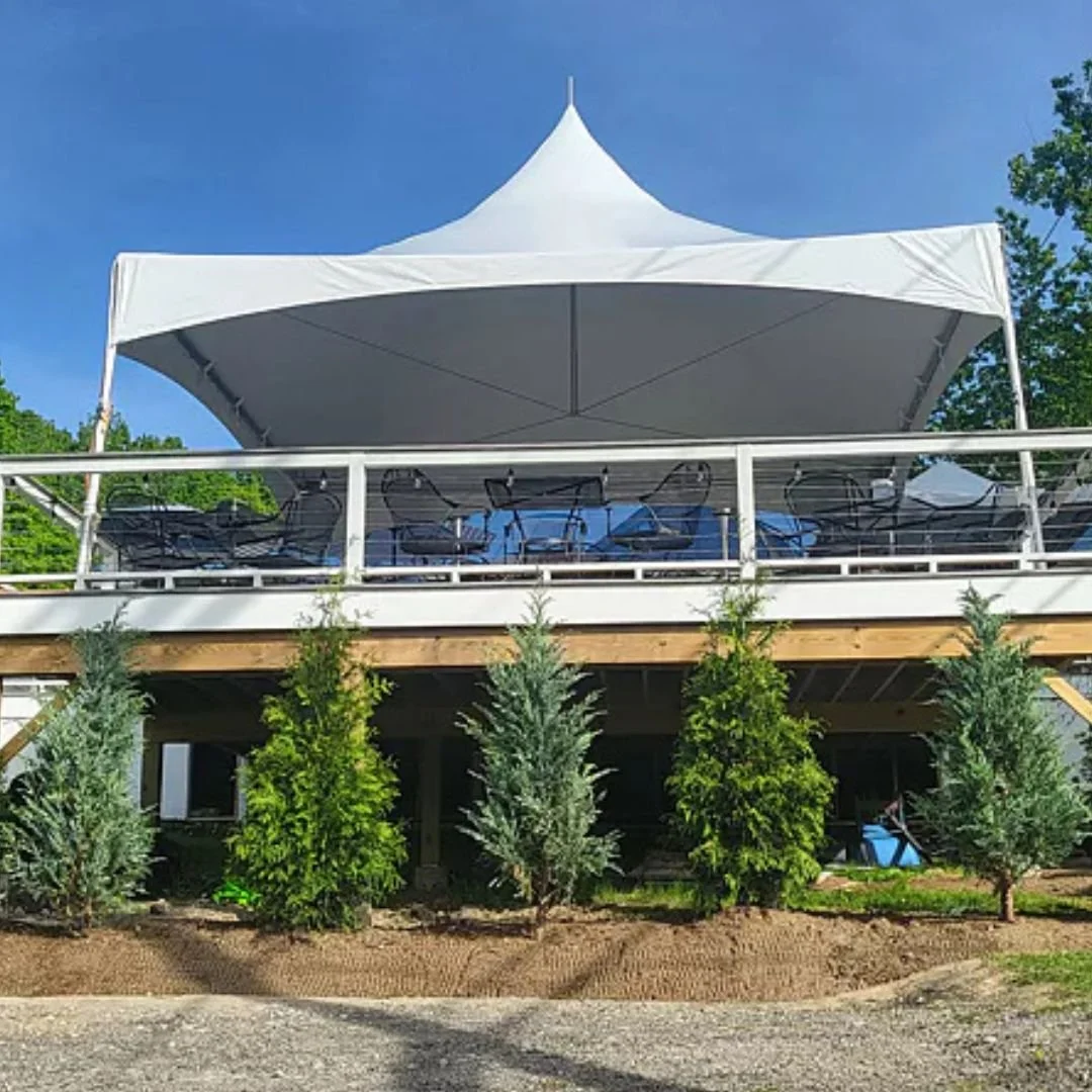 Outdoor view of a two-story structure with a white canopy and patio furniture on the upper deck, and green trees and plants on the ground level.