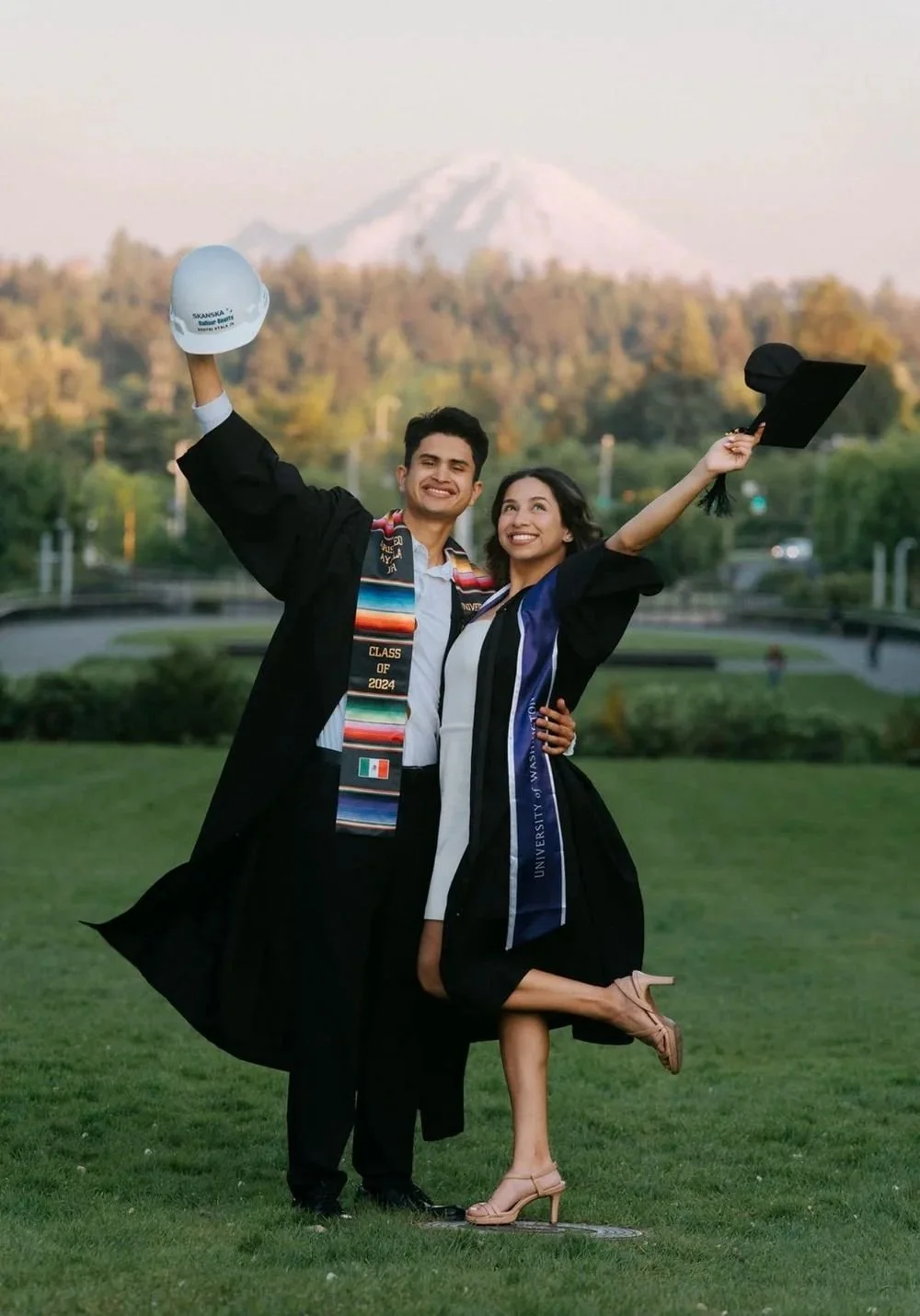 Two graduates in caps and gowns celebrating outdoors with a mountain in the background. One is holding a cap in the air, and both are smiling.