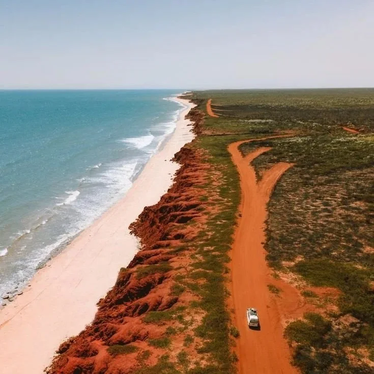 Aerial view of a coastline with a sandy beach on the left, reddish cliffs, and a dirt road running parallel to the shore on the right, with a white vehicle on the road.