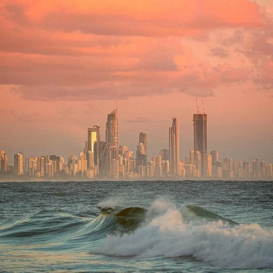 A city skyline with tall skyscrapers in the distance, viewed across a body of water with small waves during sunset or sunrise.