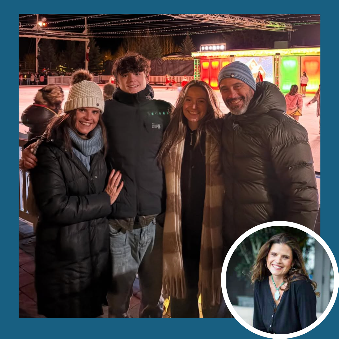 A group of four people at an ice skating rink at night, smiling and bundled in winter clothing with colorful lights and skaters in the background.