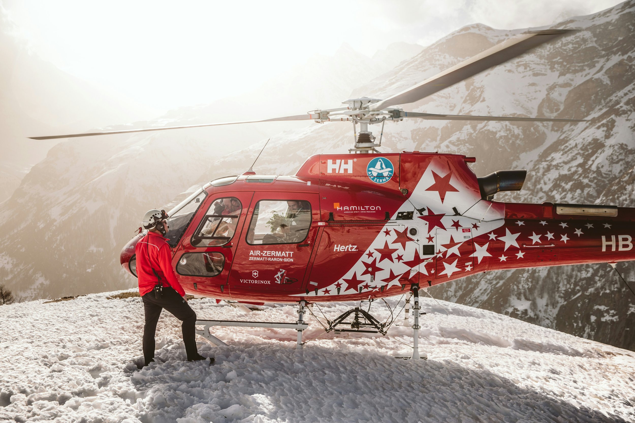 A red helicopter with a star pattern parked on snow-covered ground in a mountainous area with snow and rocky peaks in the background. Two crew members are near the helicopter, one sitting inside and two others nearby, one standing and one in a red jacket and helmet.