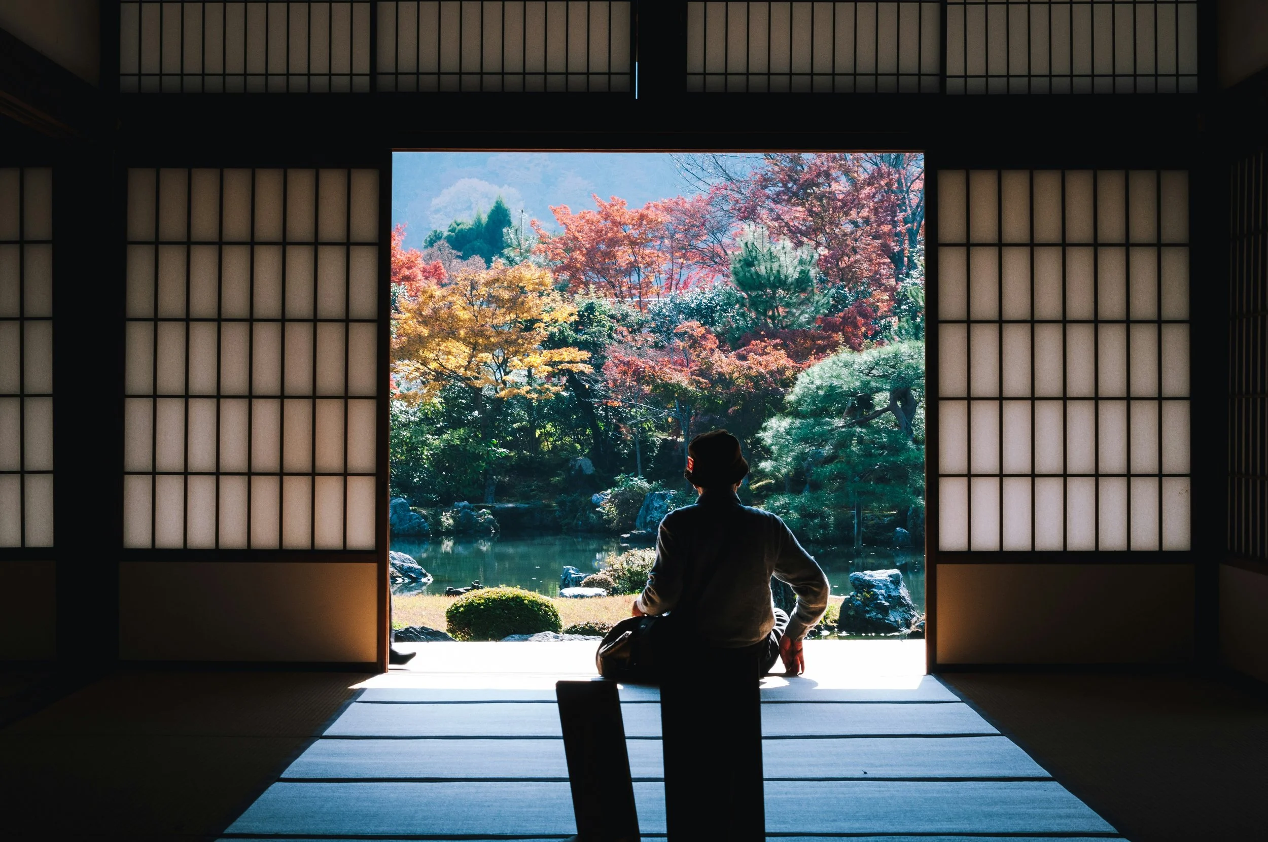 A person sitting in a traditional Japanese room, looking out through shoji doors at a garden with colorful autumn trees and a pond.
