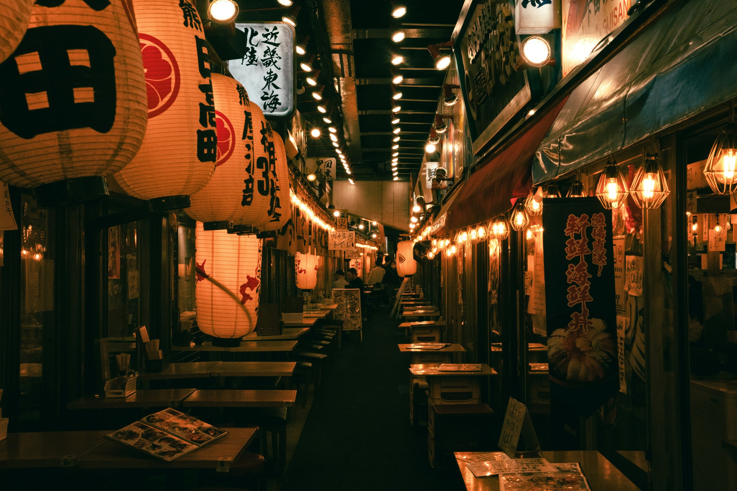 Night view of a Japanese alley adorned with traditional lanterns, illuminated signs, and restaurant lights, with seating and menus visible.