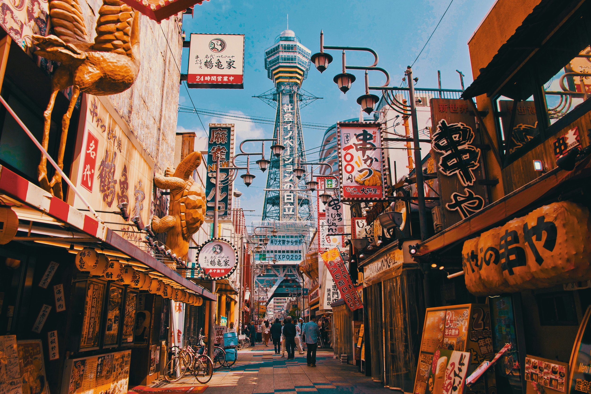 A bustling street in Japan with signs in Japanese, decorative lanterns, and a view of the Tsutenkaku Tower in the background under a blue sky.