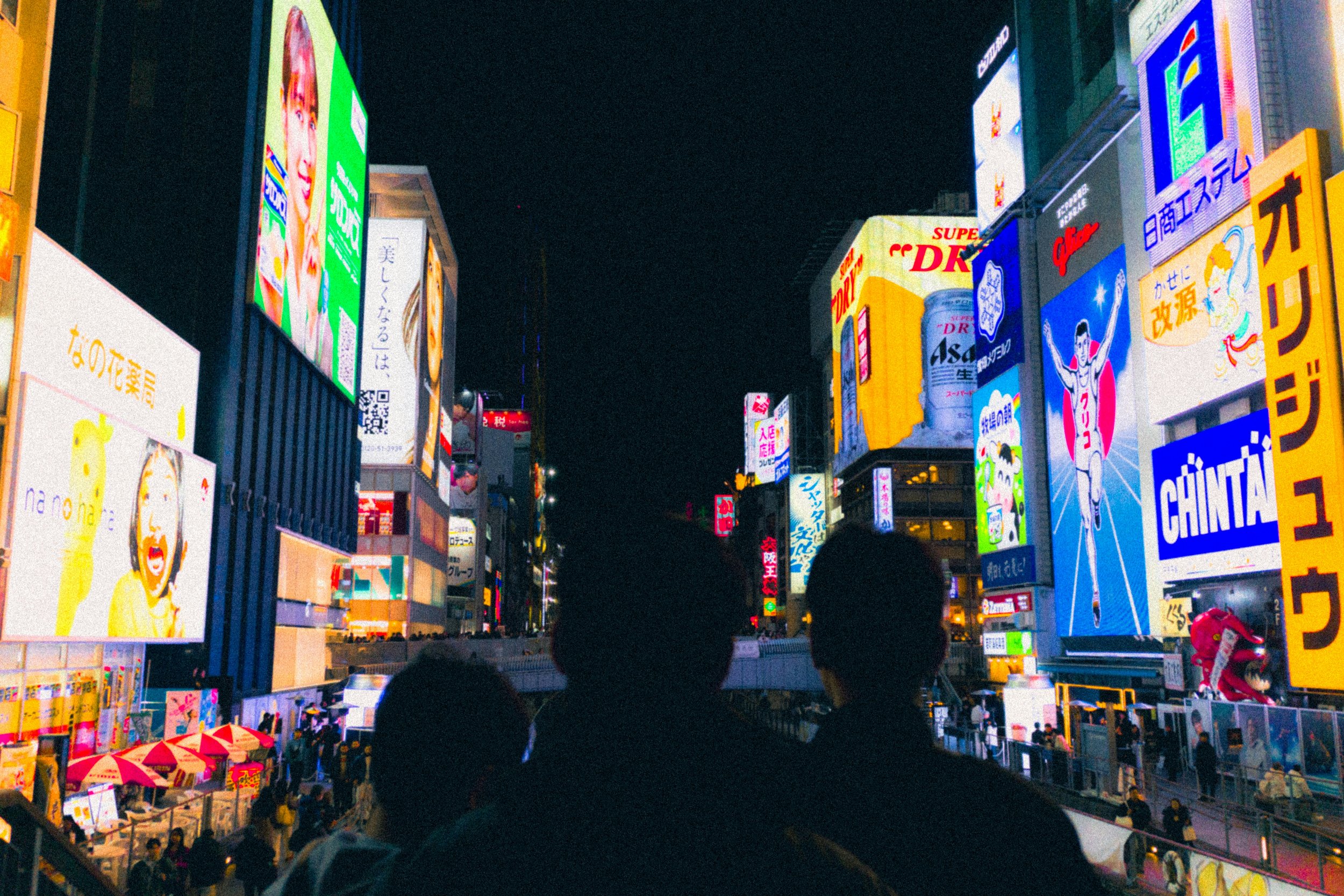 Night view of a busy street filled with bright, colorful advertisement billboards and neon signs in Japanese characters, with silhouette of people's heads in the foreground.