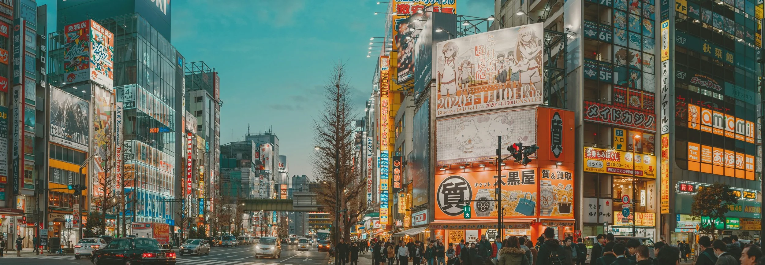 City street scene in Japan with tall buildings covered in colorful advertisements, billboards, and neon signs. Pedestrians walk on the sidewalks, with cars on the street, in front of bright, multi-story buildings on a clear evening.