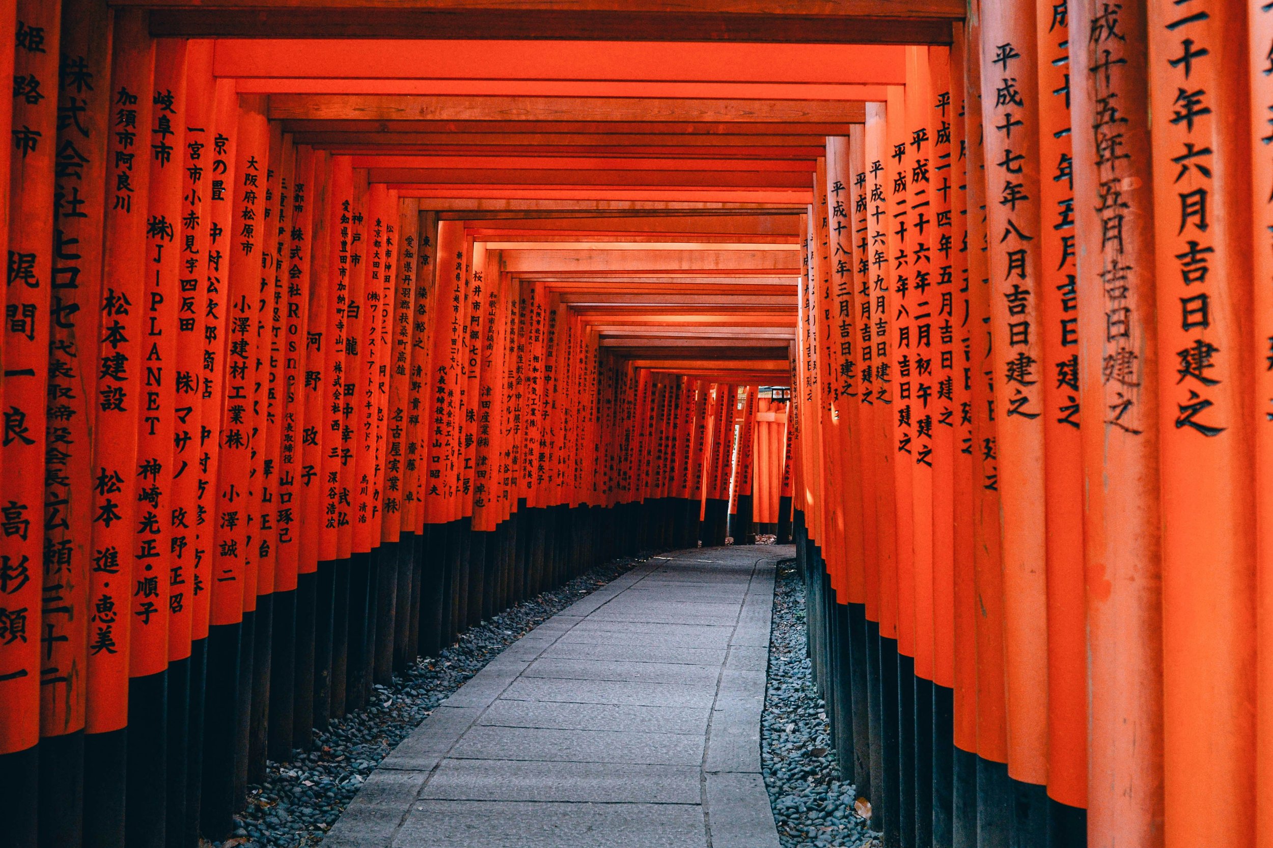 A pathway lined with numerous bright orange torii gates with black Japanese characters on them, forming an archway at Fushimi Inari Shrine in Kyoto, Japan.