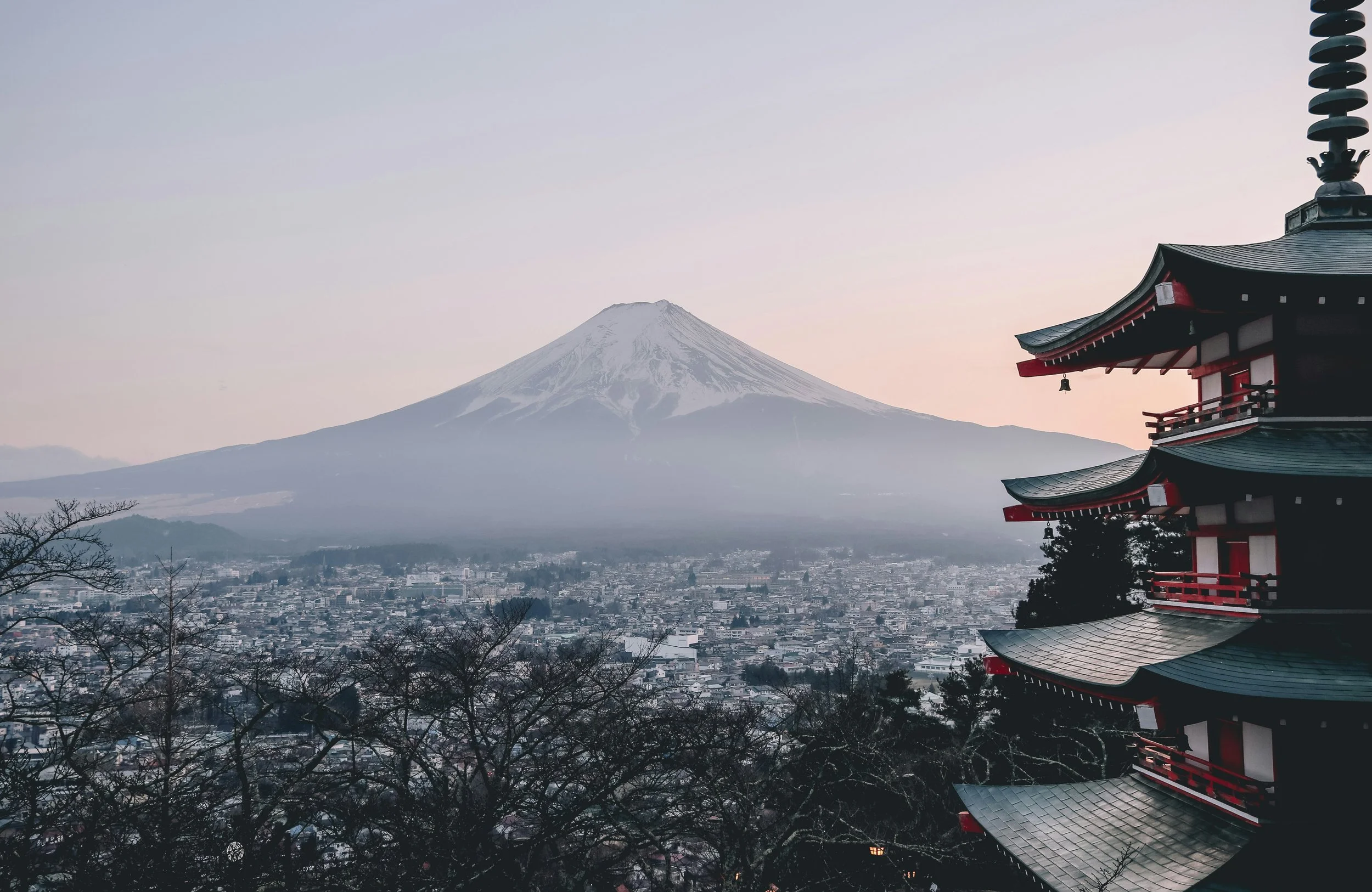 A view of Mount Fuji with snow on its peak, seen from a cityscape. A traditional Japanese pagoda with red and black details is on the right side of the image, overlooking the city and the mountain.