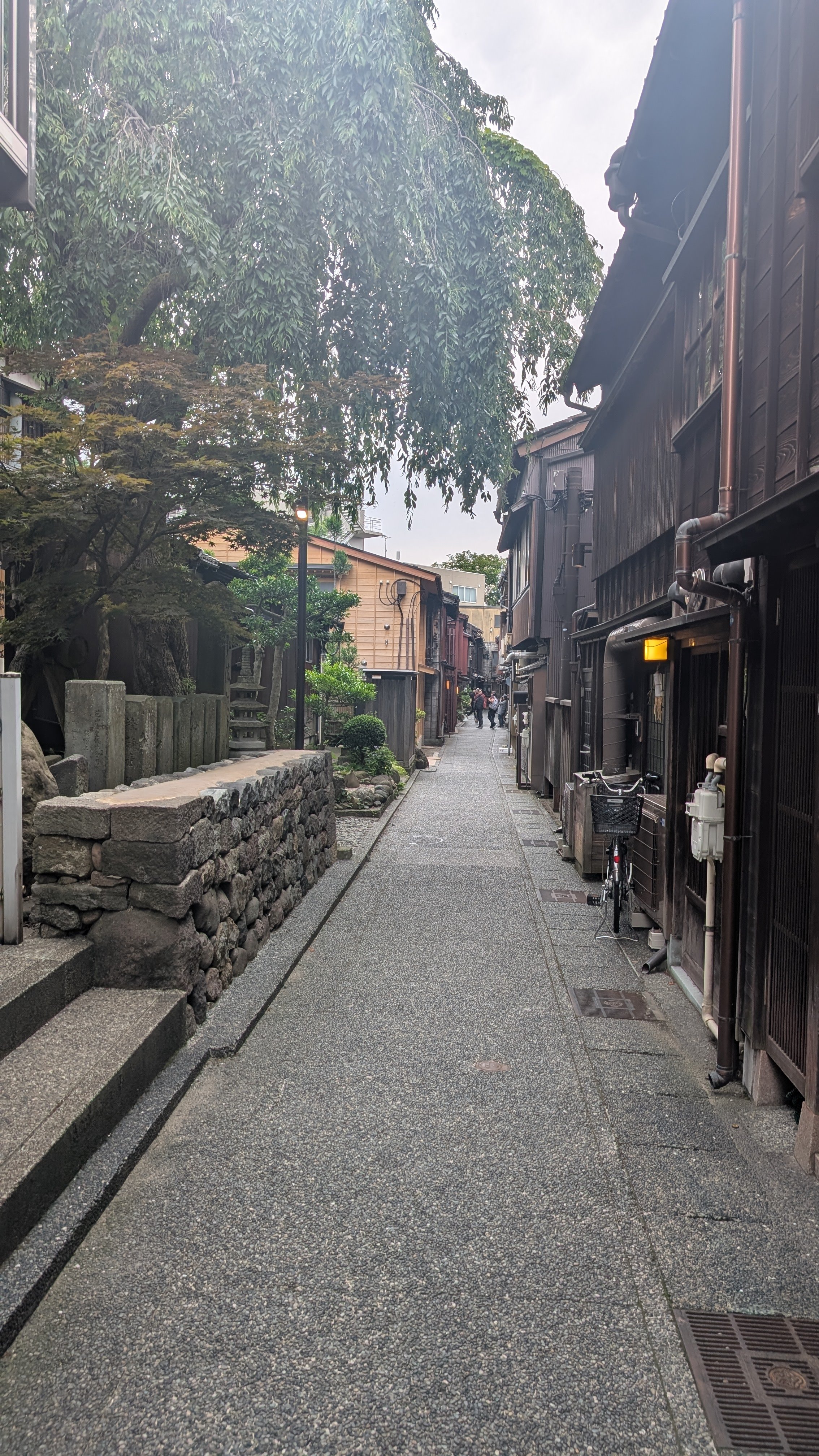 A narrow alleyway with traditional wooden buildings on the right side and a stone wall with trees on the left, with a bicycle parked near the buildings and a few people in the distance.