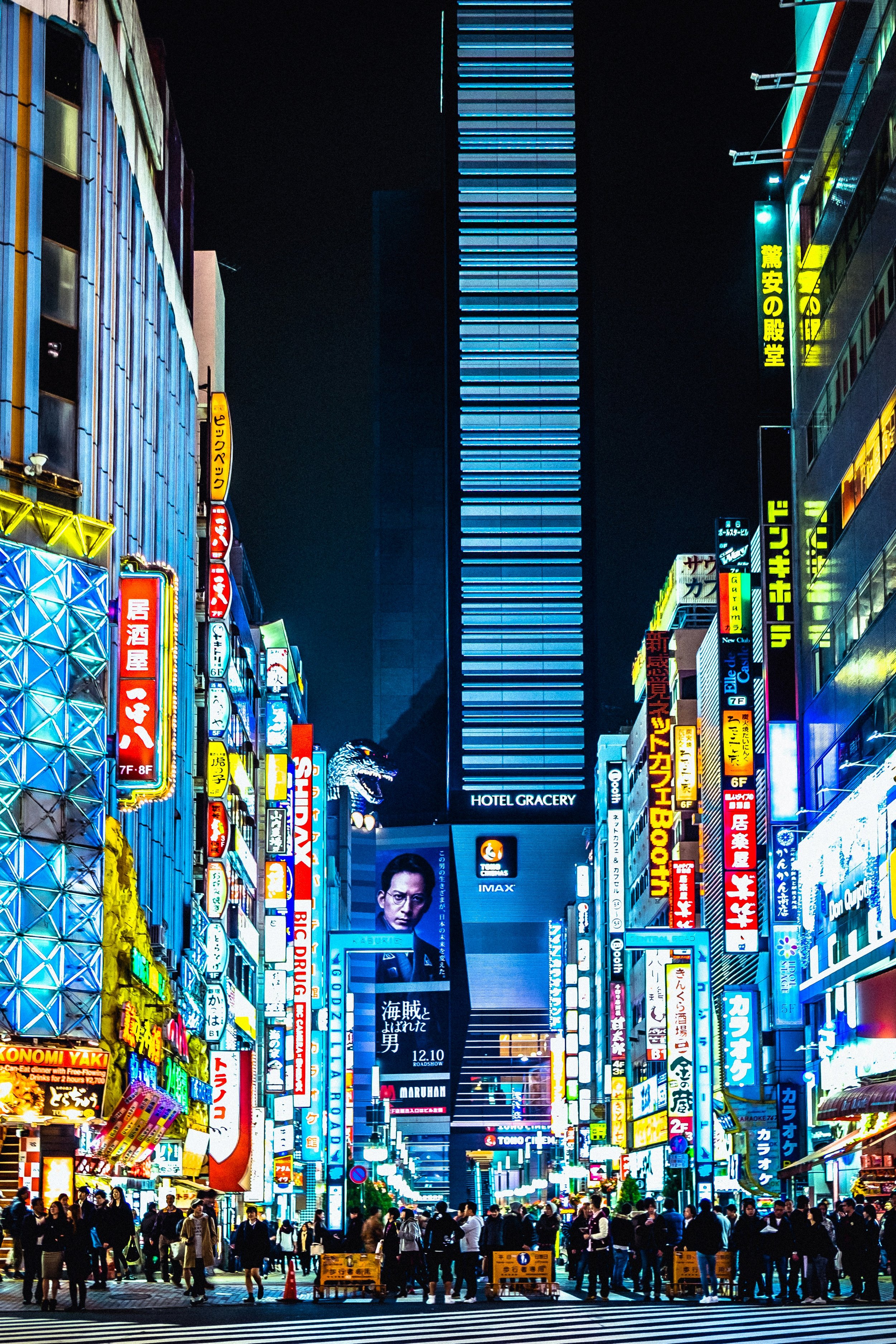 Night scene of a busy commercial street with brightly lit signs and advertisements in various languages, with a crowd of people crossing at a crosswalk.