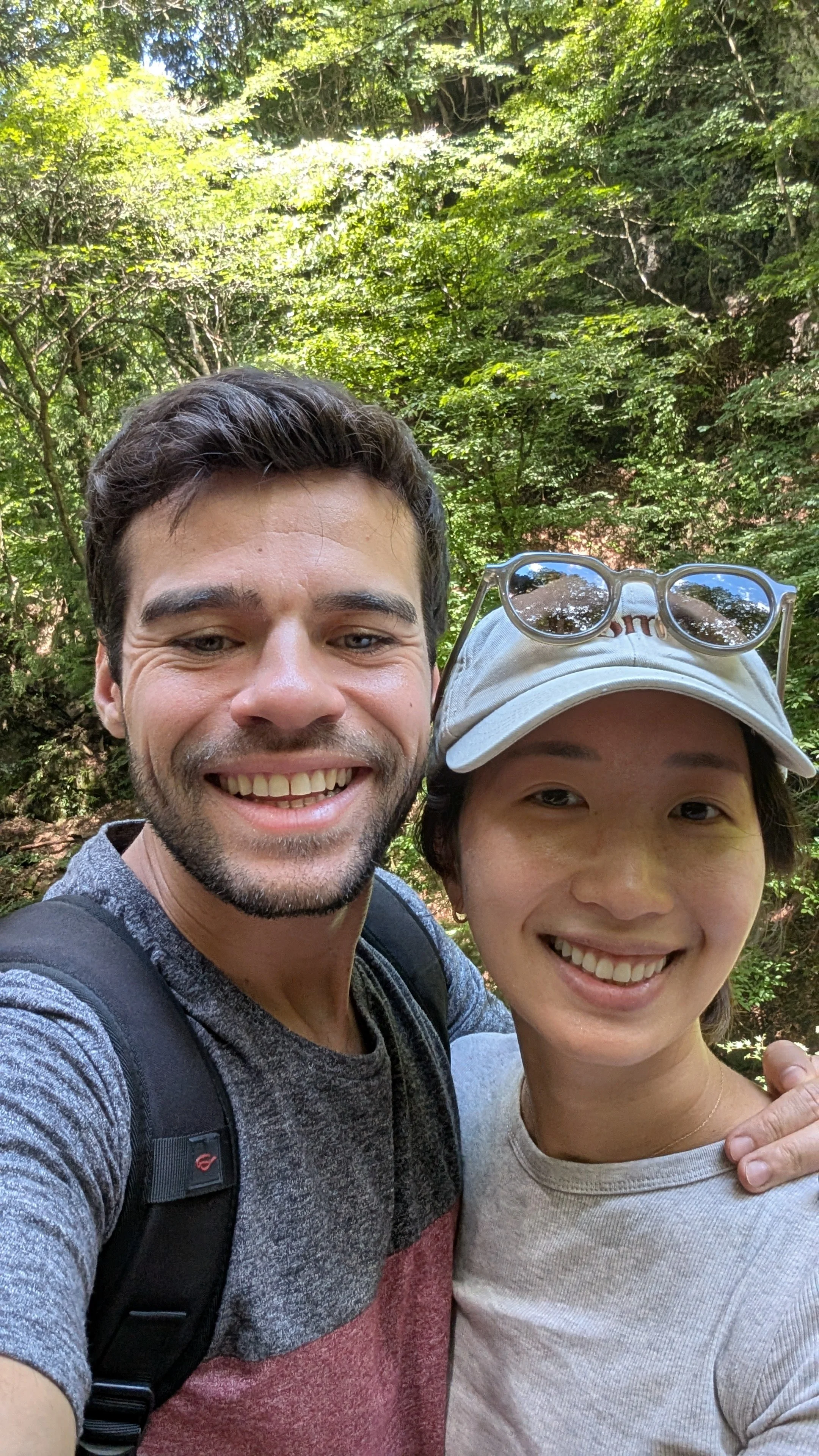Two people taking a selfie in a lush green forest. The man on the left has short dark hair, facial hair, and is smiling. He is wearing a gray and red shirt and a black backpack. The woman on the right has short dark hair, is wearing sunglasses on her hat, and a light-colored top. She is also smiling. They are close together, enjoying nature.