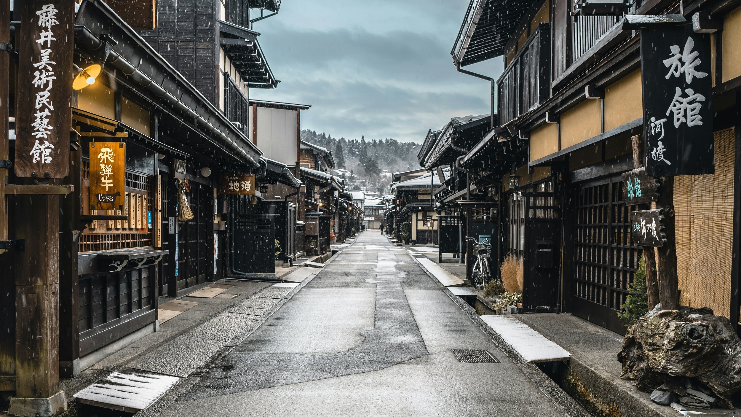 A quiet, rainy, traditional Japanese street with wooden buildings on both sides, some signs with Japanese characters, and a bicycle parked on the right side; mountainous landscape visible in the background.