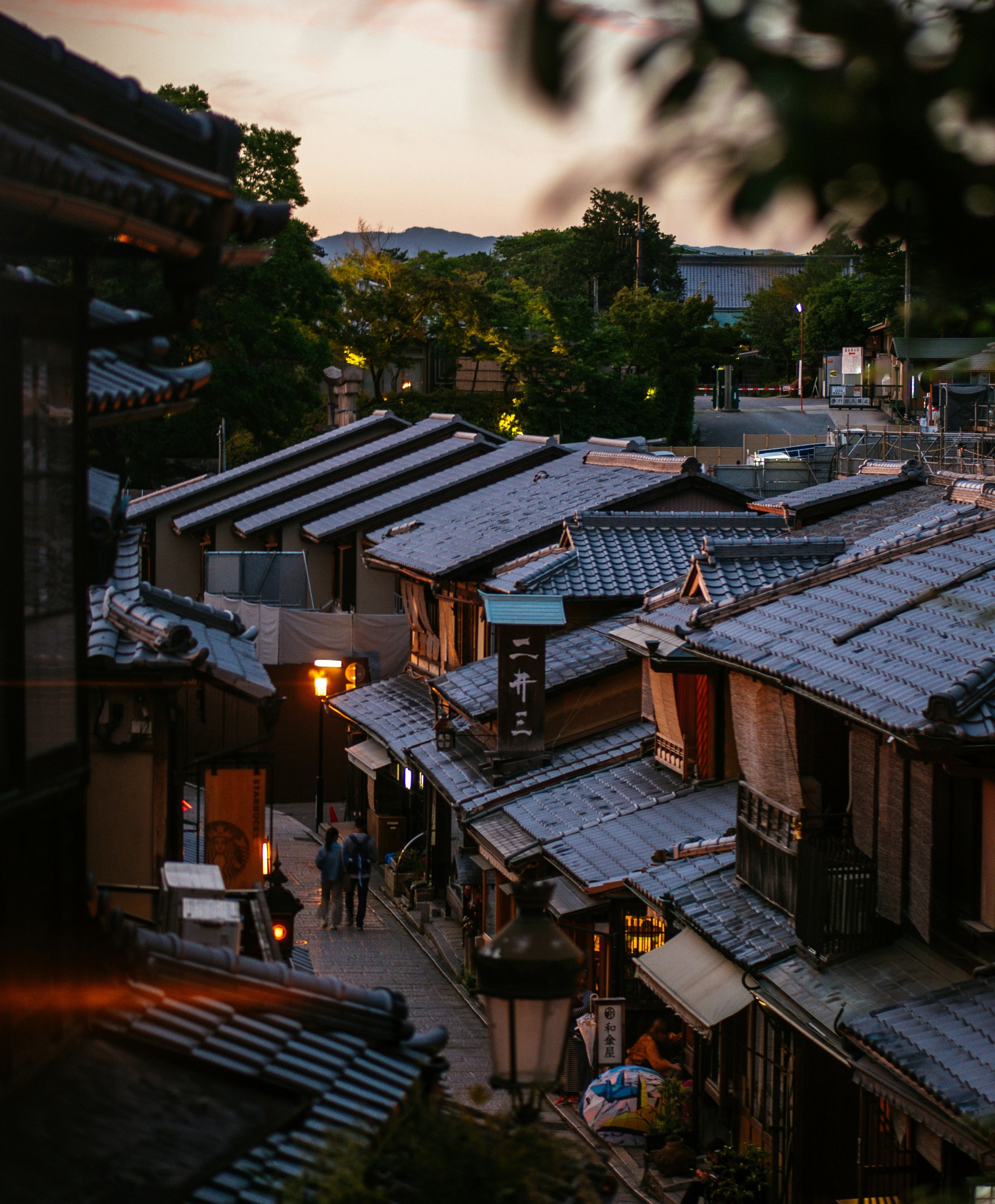 A traditional Japanese street scene during dusk with tiled rooftops, narrow alleyway, some shops, trees, and distant mountains in the background.