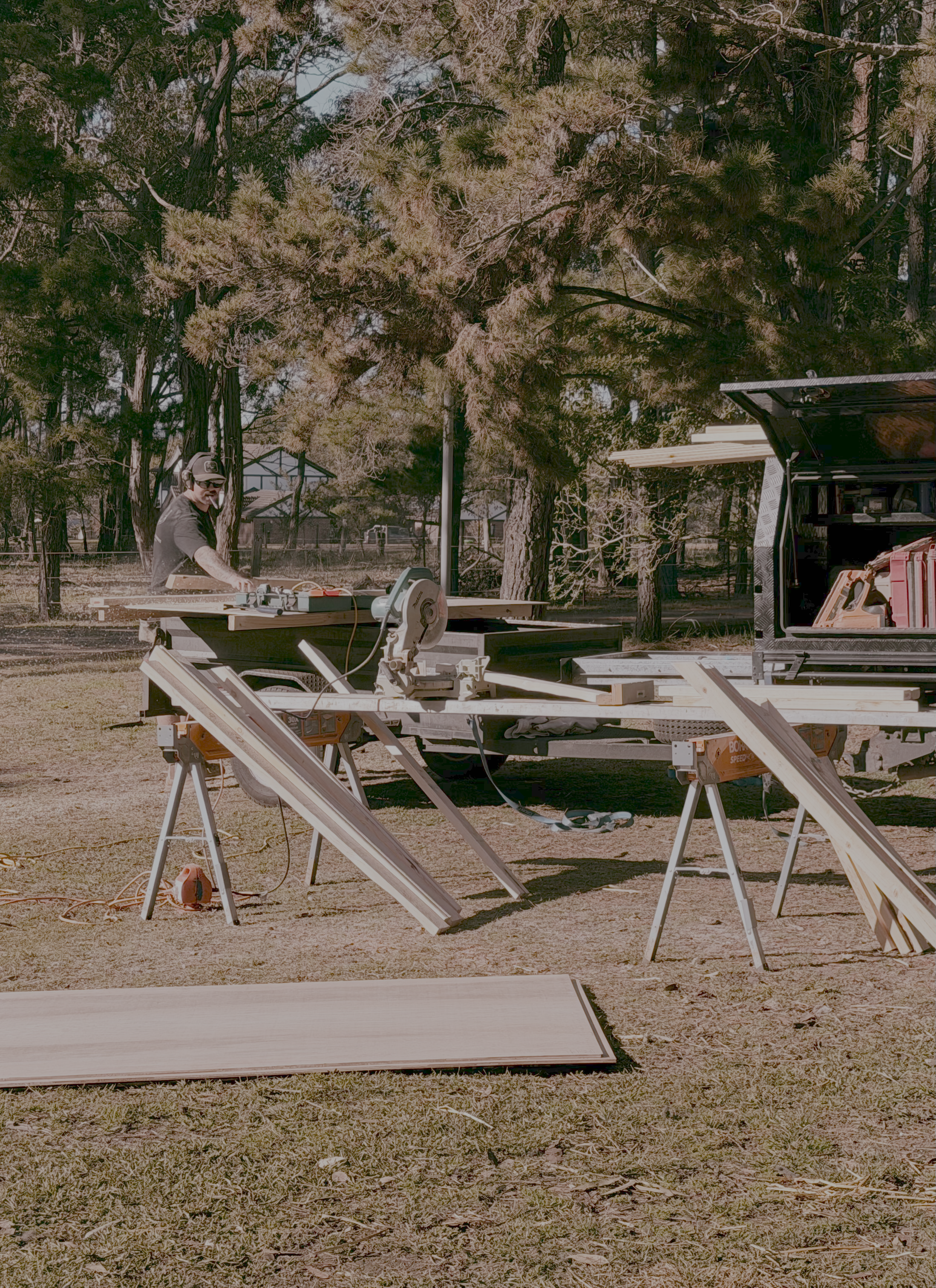 A man working outdoors with several saws and wood planks, with tall trees and a truck in the background.