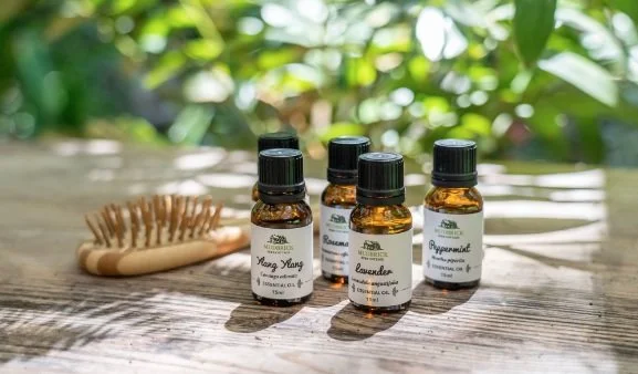Five small amber glass bottles of essential oils labeled with different scents, placed on a wooden surface with a natural outdoor background, and a wooden massage brush beside them.