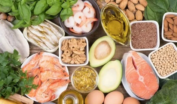 Ingredients including shrimp, avocado, salmon, eggs, olive oil, lentils, chickpeas, and various herbs and vegetables on a wooden surface.