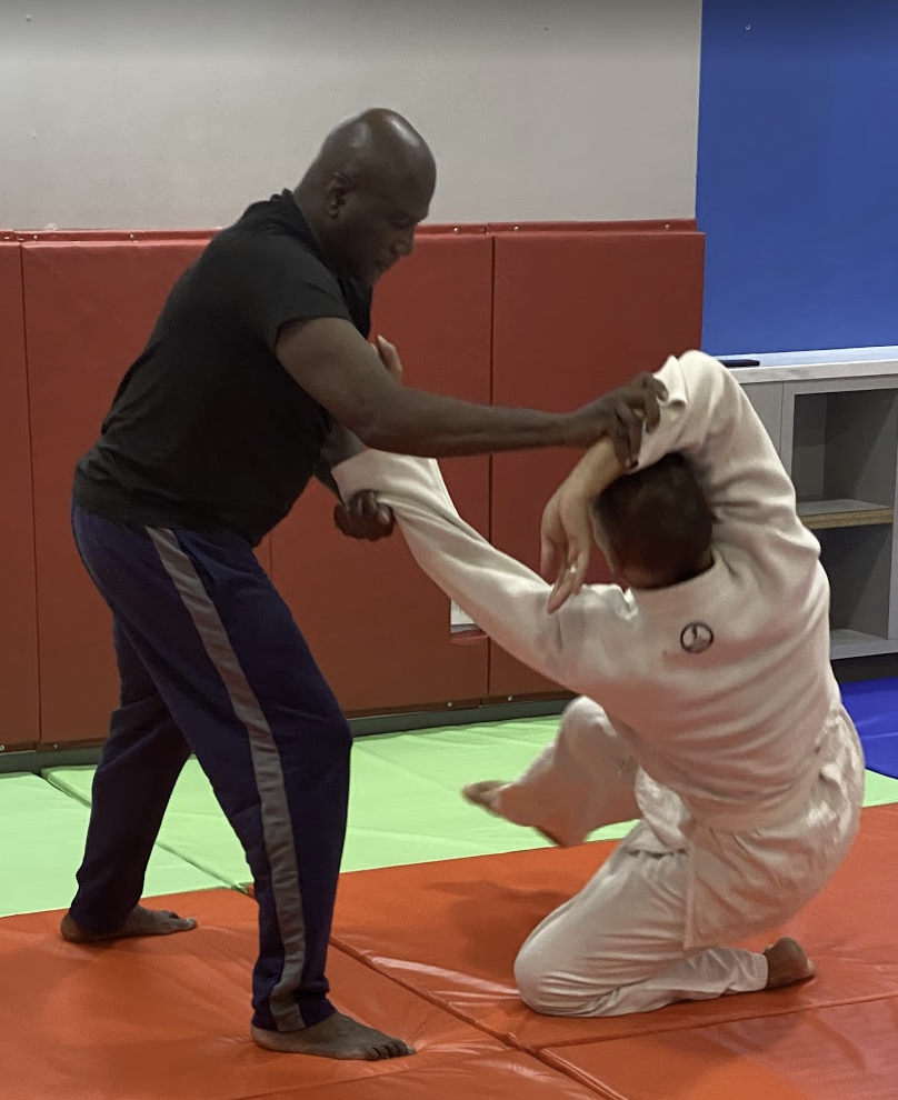 Two men practicing Brazilian Jiu-Jitsu on colorful mats; one is in a white gi, kneeling, the other in a black t-shirt and dark pants, standing and grappling.