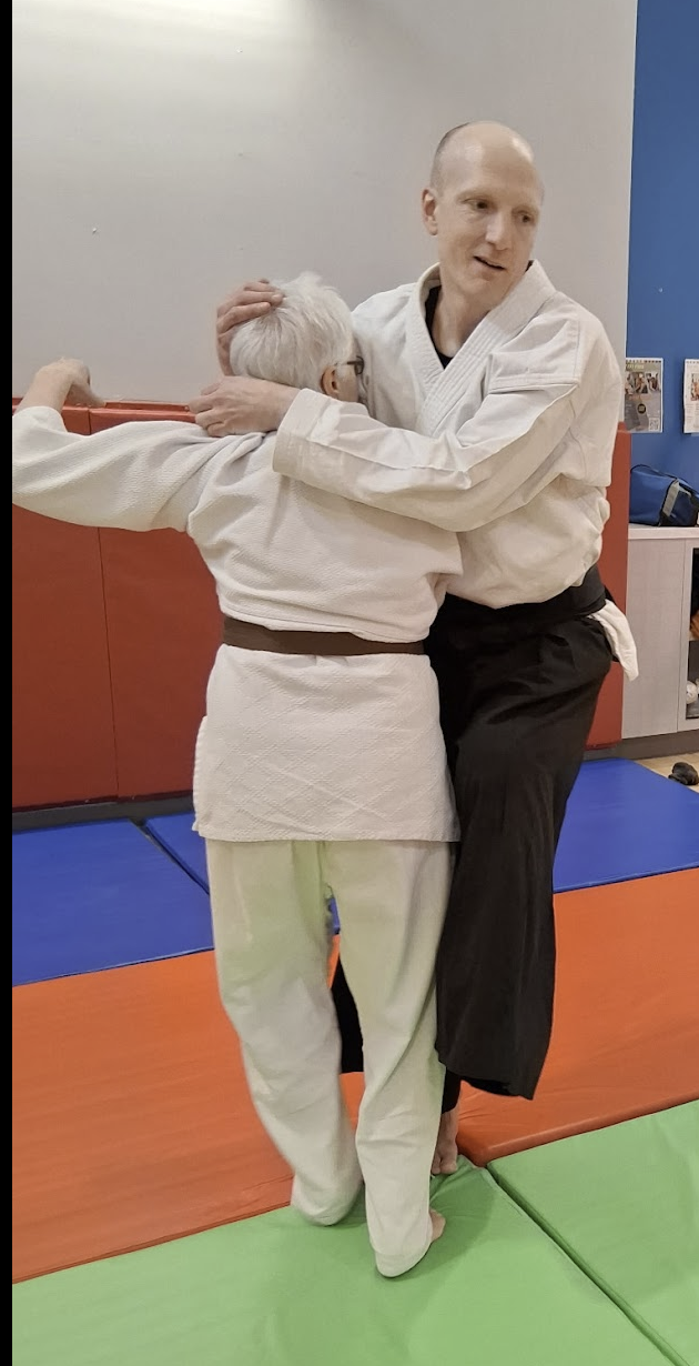 Two people practicing judo or martial arts on a colorful mat in a dojo, with one person supporting the other in a hold.