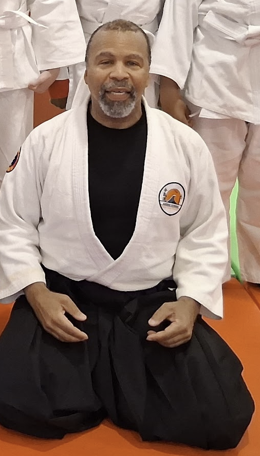 A man in a martial arts gi sitting on the floor during a group training session.