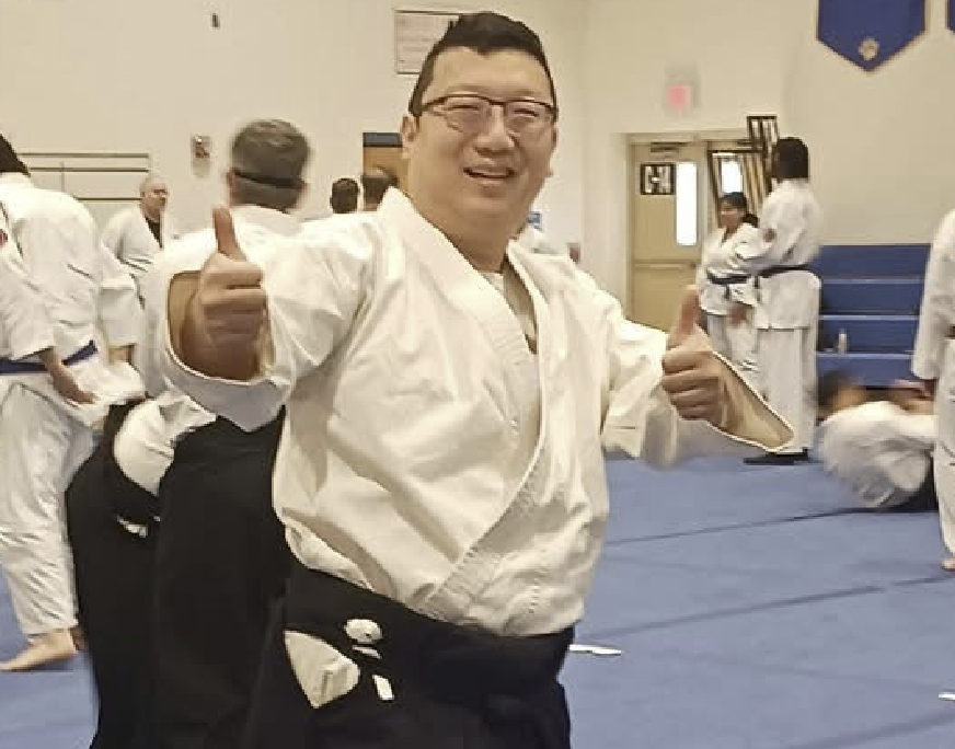 A man in a karate gi giving a thumbs-up inside a dojo with other martial artists in the background.