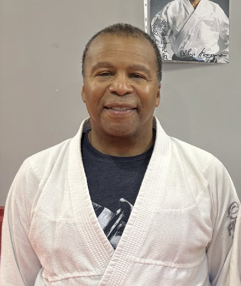 A man wearing a white martial arts gi, standing in front of a gray wall with a poster of a martial artist in a gi.