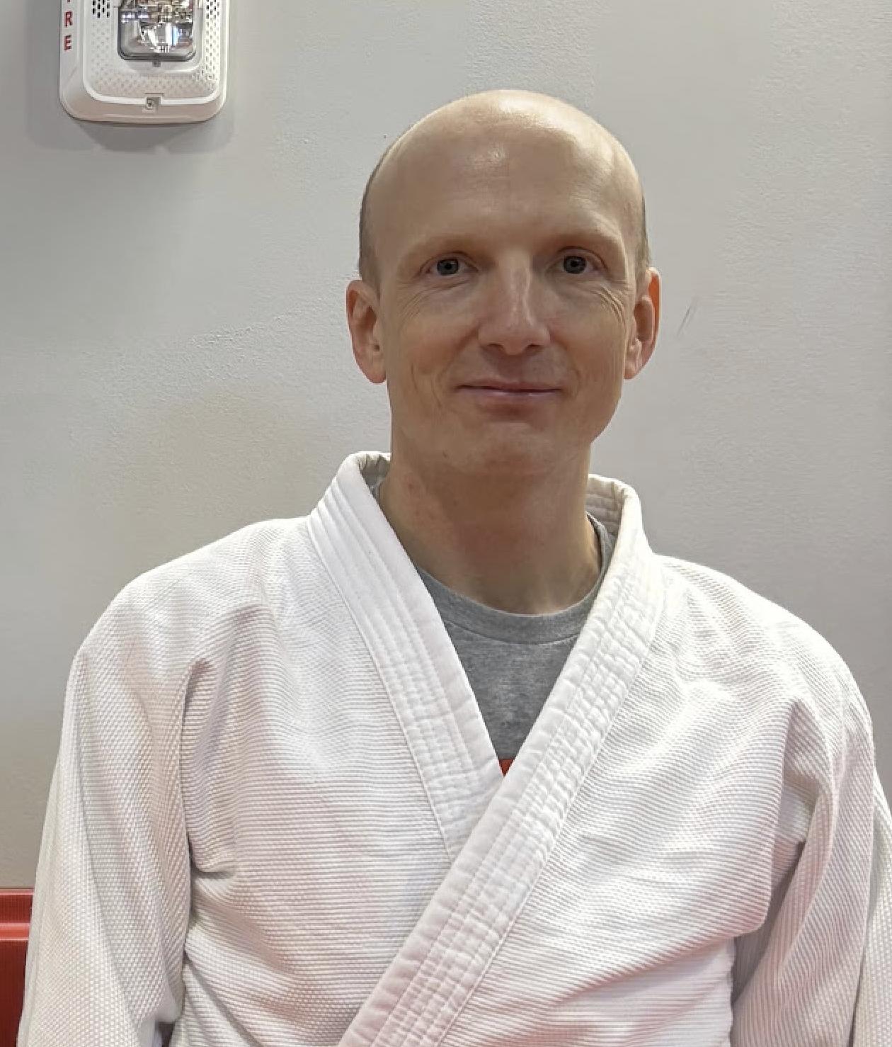 A man with a shaved head wearing a white martial arts gi uniform, standing indoors against a beige wall with a fire alarm on the upper left.