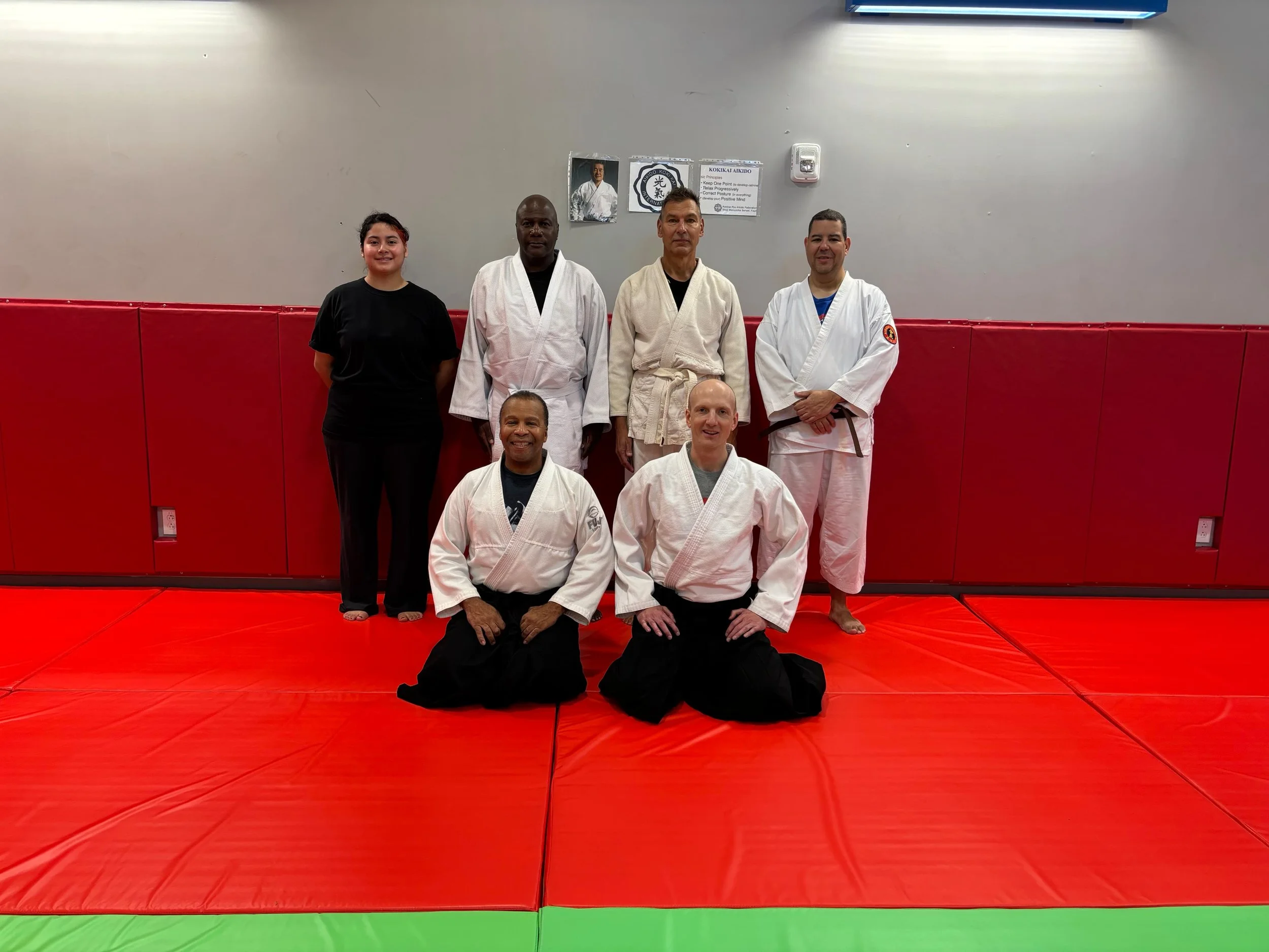 Group of six martial artists, three in white gi, one in black gi, and one woman in black shirt, posing in a dojo with red mats and a gray wall. There are posters and a speaker on the wall behind them.