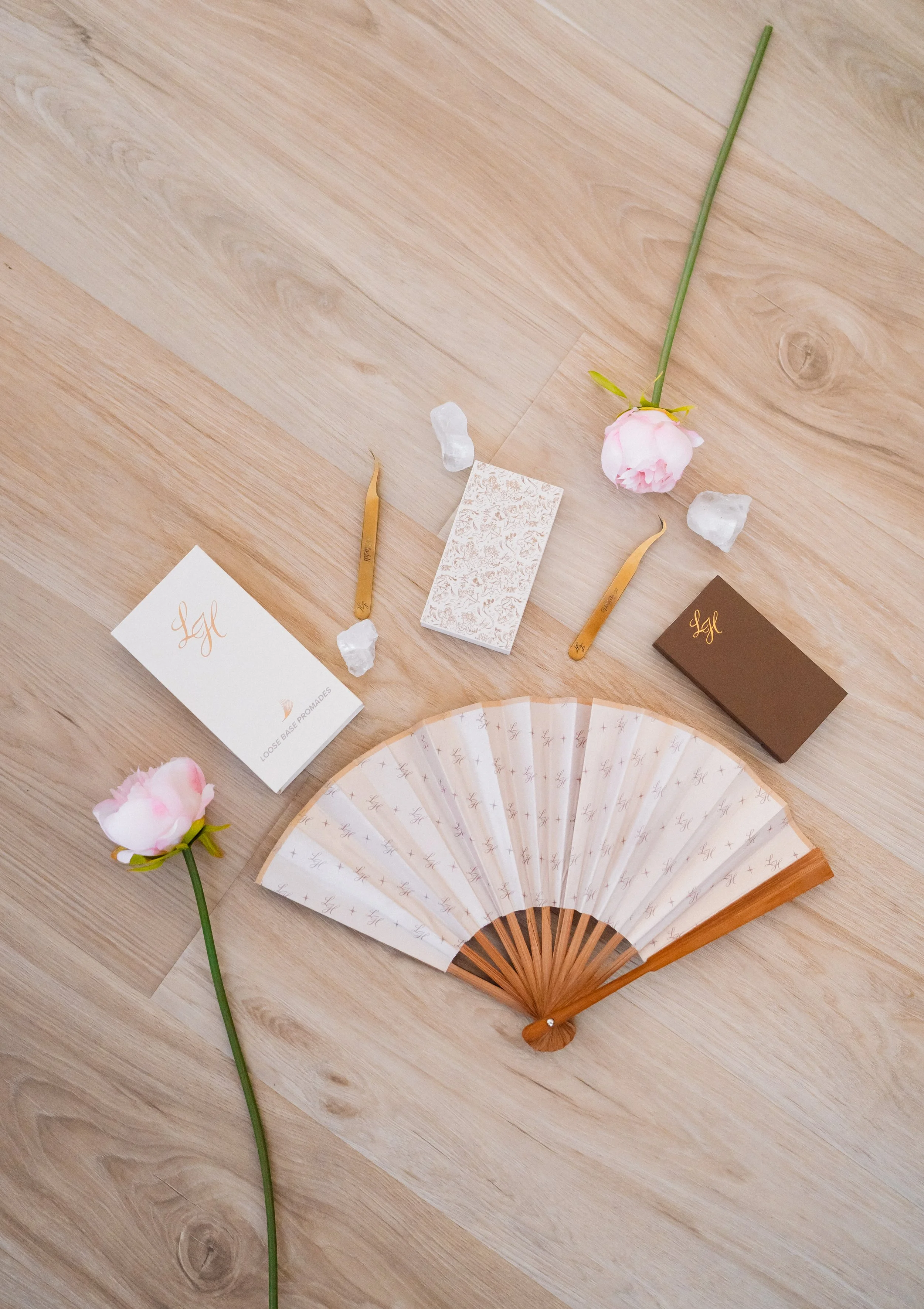 "Beauty N Artistry lash tools including gold tweezers, lash boxes, crystals, and pink roses on wood floor