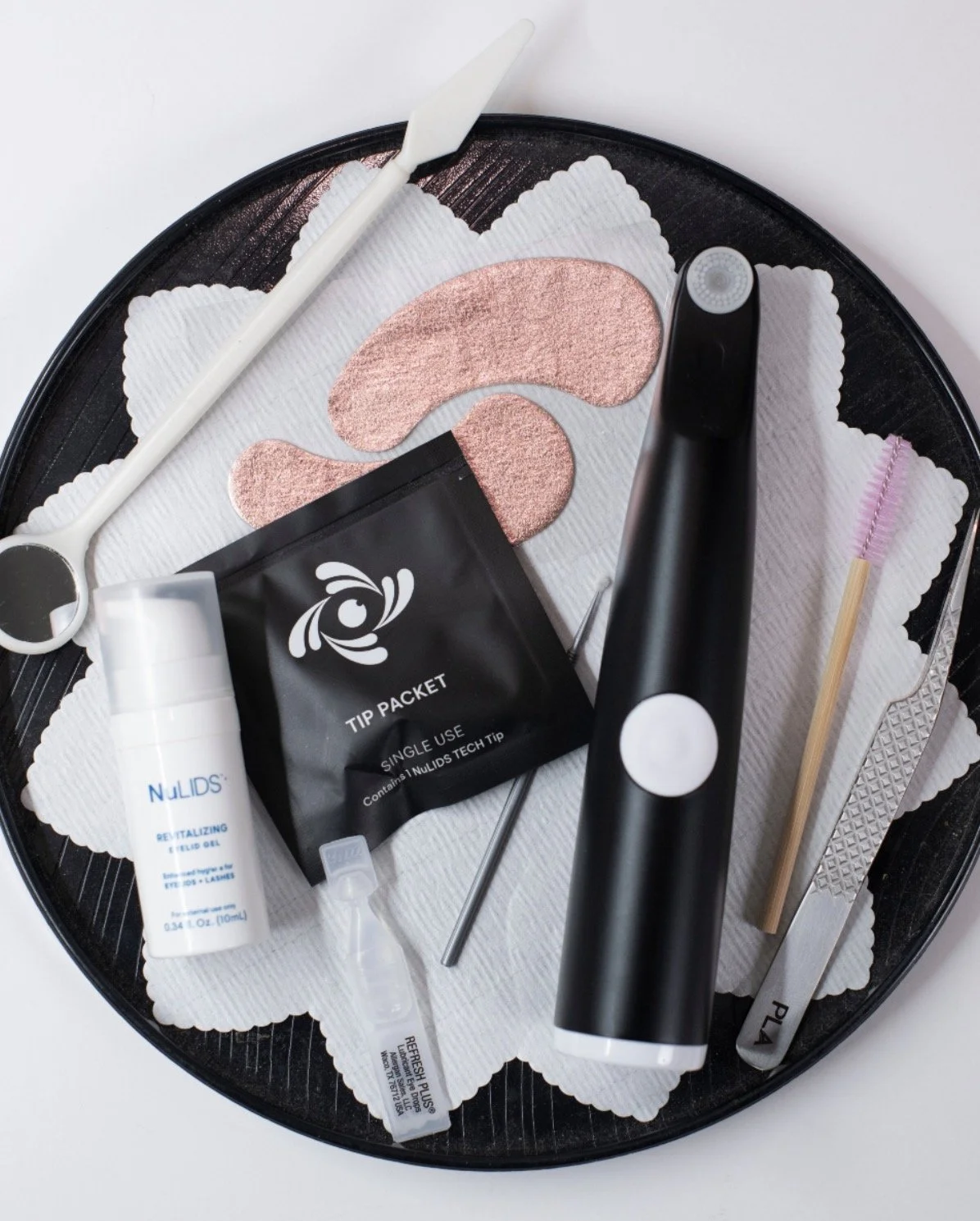 A tray with  beauty tools, including eye patches, a facial roller, a cleaning device, a tip packet, and small applicator brushes, arranged on tissue paper.